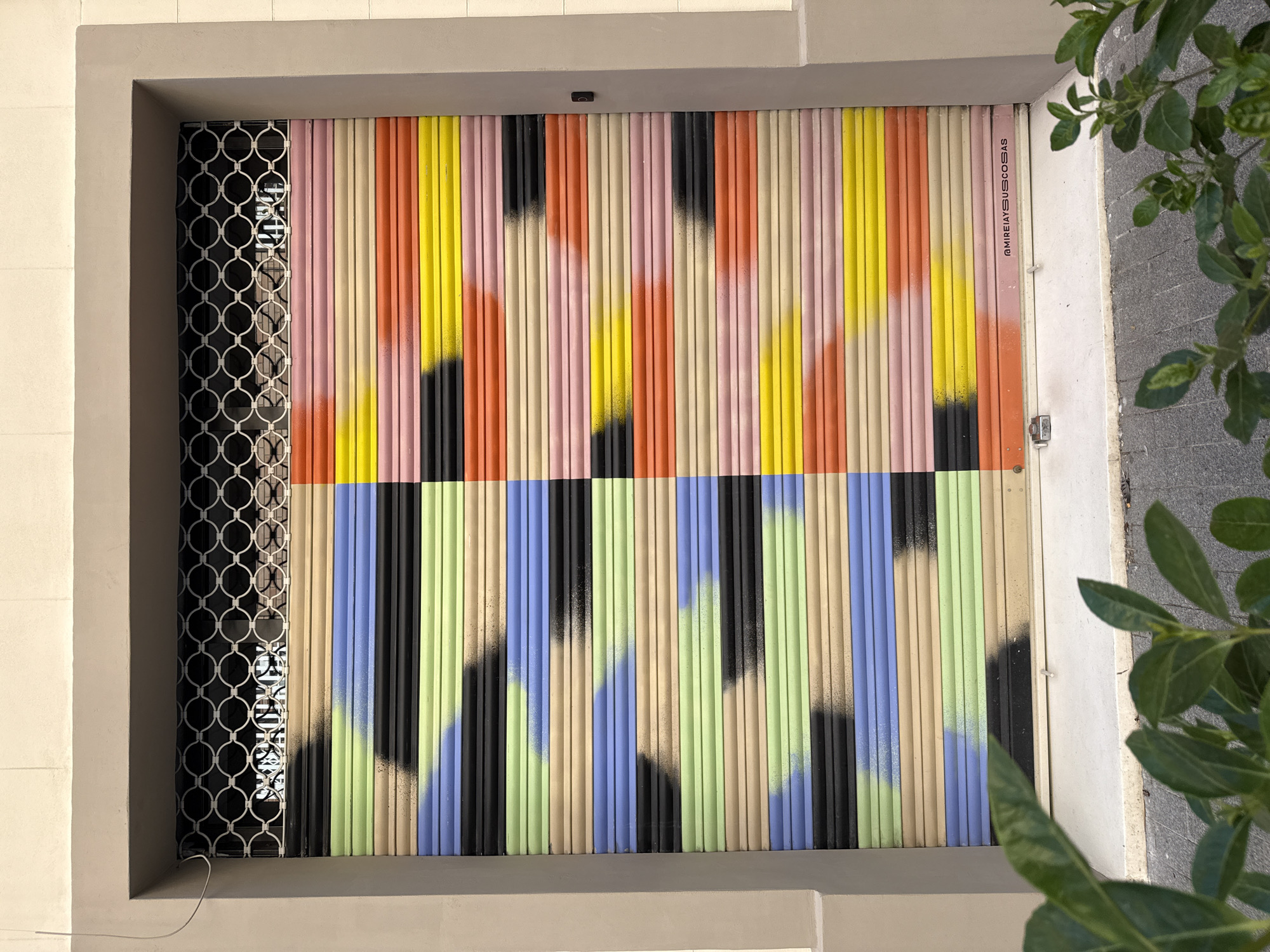 The image captures a storefront in Barcelona, Spain, featuring a corrugated metal roll-down shutter door, divided into two horizontal sections, each with distinct color schemes. The upper section has vertical stripes of yellow, orange, pink, and beige with black accents near the middle of the section, and the lower section uses colors like blue, green, beige, and black, each also accented with spray paint elements. The left side of the storefront has a black, decorative metal grate. The door is surrounded by a gray concrete frame. Green foliage partially obscures the right side of the frame. A surveillance camera is visible at the top of the door. A vertical sign on the right has the text "@MIREIAYUSUSCOSAS."