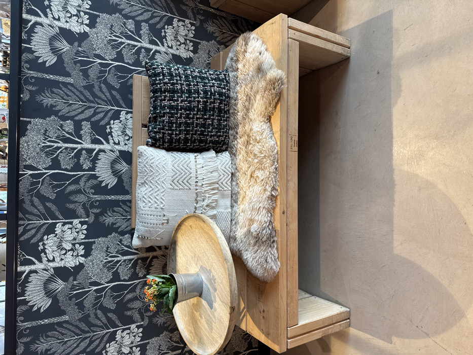 This interior scene, located in Altenheim, Germany, presents a light-colored wooden bench positioned against a wall covered in dark blue wallpaper featuring a white or silver pattern of stylized trees and foliage. The bench has a solid wooden seat and a slatted wooden backrest. On the bench, from left to right, a round wooden tray holds a small galvanized metal pot containing a green plant with numerous small orange flowers. To its right, an off-white decorative pillow with geometric patterns, fringe, and metallic embellishments rests against the backrest. Adjacent to it, a dark green and black woven tweed-like pillow is also positioned. A light brown faux fur rug or throw is draped across the bench seat, extending partially beneath the pillows. The floor is a light-colored, smooth surface, possibly concrete, exhibiting shadows cast by the bench and its contents. No individuals are present, and no actions are observed within the frame.