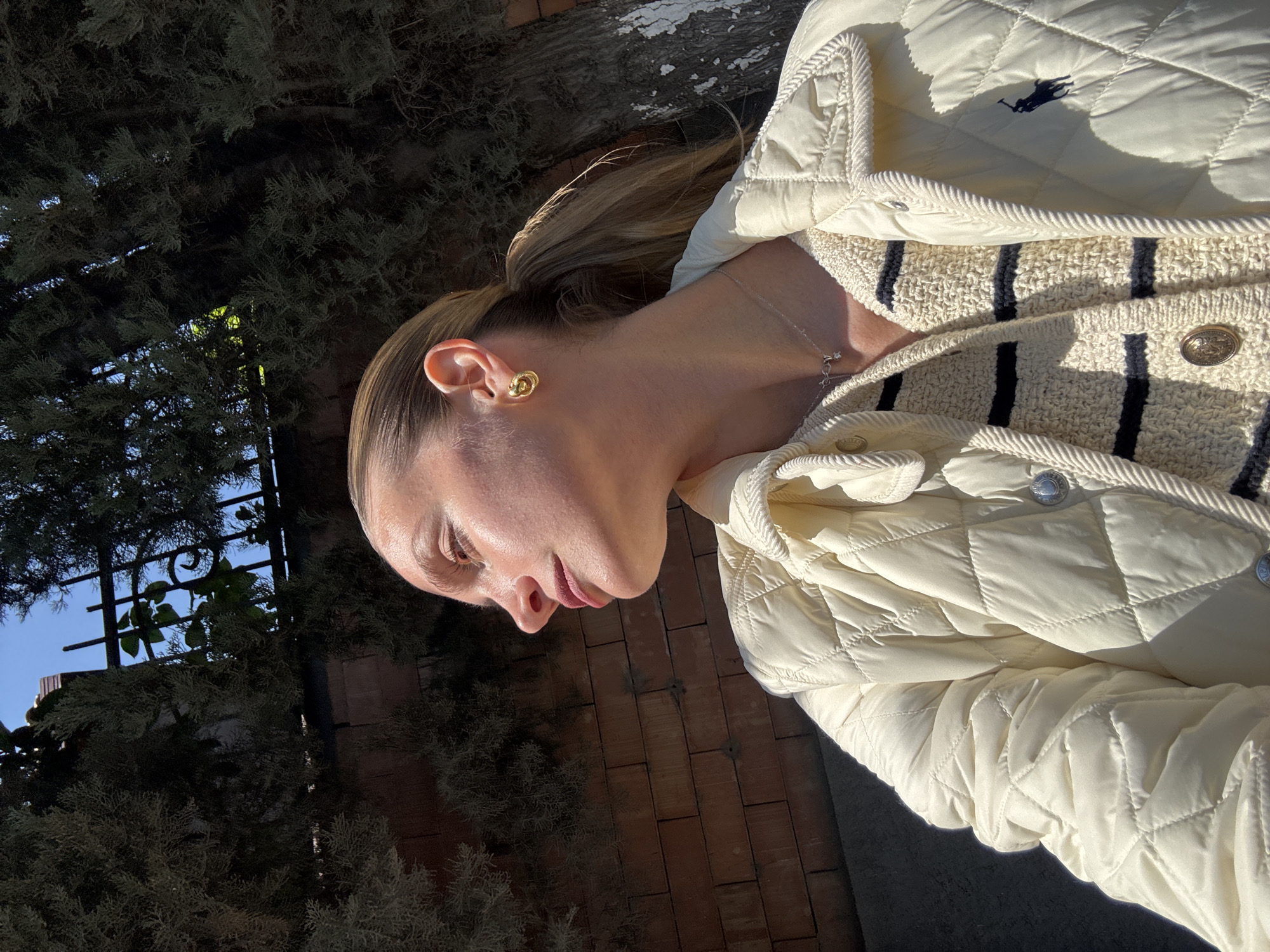 A woman in Unknown, Unknown is outdoors, near a brick wall and some dense, dark green bushes. She is wearing a light-colored, quilted Ralph Lauren jacket over a striped top with gold buttons. The woman is wearing gold earrings and a delicate necklace. Her hair is pulled back in a ponytail.