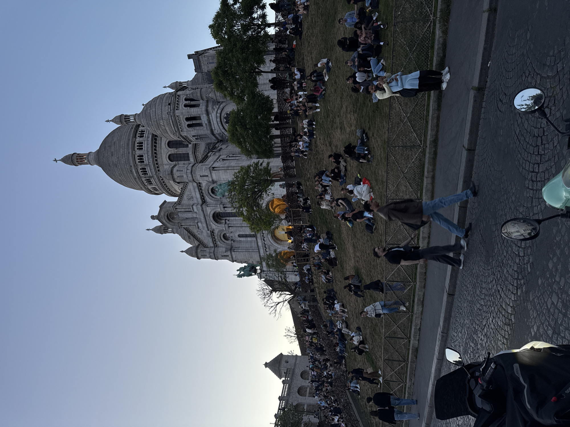 The image, taken in Montmartre, Paris, depicts a serene early evening scene with the iconic Sacré-Cœur Basilica prominently on the left. The sky is a clear, light blue, indicative of the golden hour or late afternoon.
In the foreground, a paved cobblestone street runs along the right side of the frame, where parts of a dark scooter or motorcycle are visible, including its handlebars and two rearview mirrors. One mirror reflects distant buildings, while a faint "SYNERGY" can be seen on the scooter's handlebar cover.
Separating the street from the basilica is a grassy slope, densely populated with numerous people. They are sitting or lying down, facing generally towards the basilica or enjoying the expansive view, engaging in conversation, and soaking in the relaxed atmosphere. A metal fence borders the grassy area. Several individuals are also seen walking along the paved path.
The Sacré-Cœur Basilica itself, with its distinctive white domes, ornate spires, and intricate architectural details, stands majestically. The overall ambiance is one of leisure and communal enjoyment, with people gathered to appreciate the setting and scenery under pleasant weather conditions. Trees are scattered throughout the landscape, adding greenery to the urban environment.