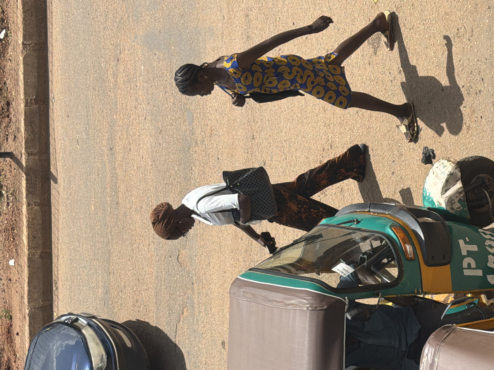 Two young women are walking on a dusty, paved road in what appears to be a rural or semi-urban setting in Bukuru, Nigeria. The sun is shining brightly, casting long shadows, indicating it's likely midday or early afternoon.
The woman in the foreground, on the right, is wearing a white t-shirt, dark pants with a rust-colored pattern, and a brown headscarf. She carries a black and grey checkered bag over her shoulder and holds a dark-colored mobile phone in her right hand. She is looking down.
The second woman, further up the road and to the left, is wearing a brightly colored yellow and blue patterned dress. She has her hair braided and is wearing earrings. She carries a black bag strapped across her body. Her gaze is directed forward.
A distinctive three-wheeled auto-rickshaw, painted green, yellow, and white, is parked or moving slowly on the right side of the frame, partially obscuring the road. The text "PT-1508" is visible in white on the side of the vehicle. In the bottom left corner of the image, the top of a dark blue and black suitcase or bag is visible. The ground is a mix of asphalt and dirt, with a low, overgrown border on the left side of the image, suggesting a roadside location. The overall impression is one of everyday life in a Nigerian town.