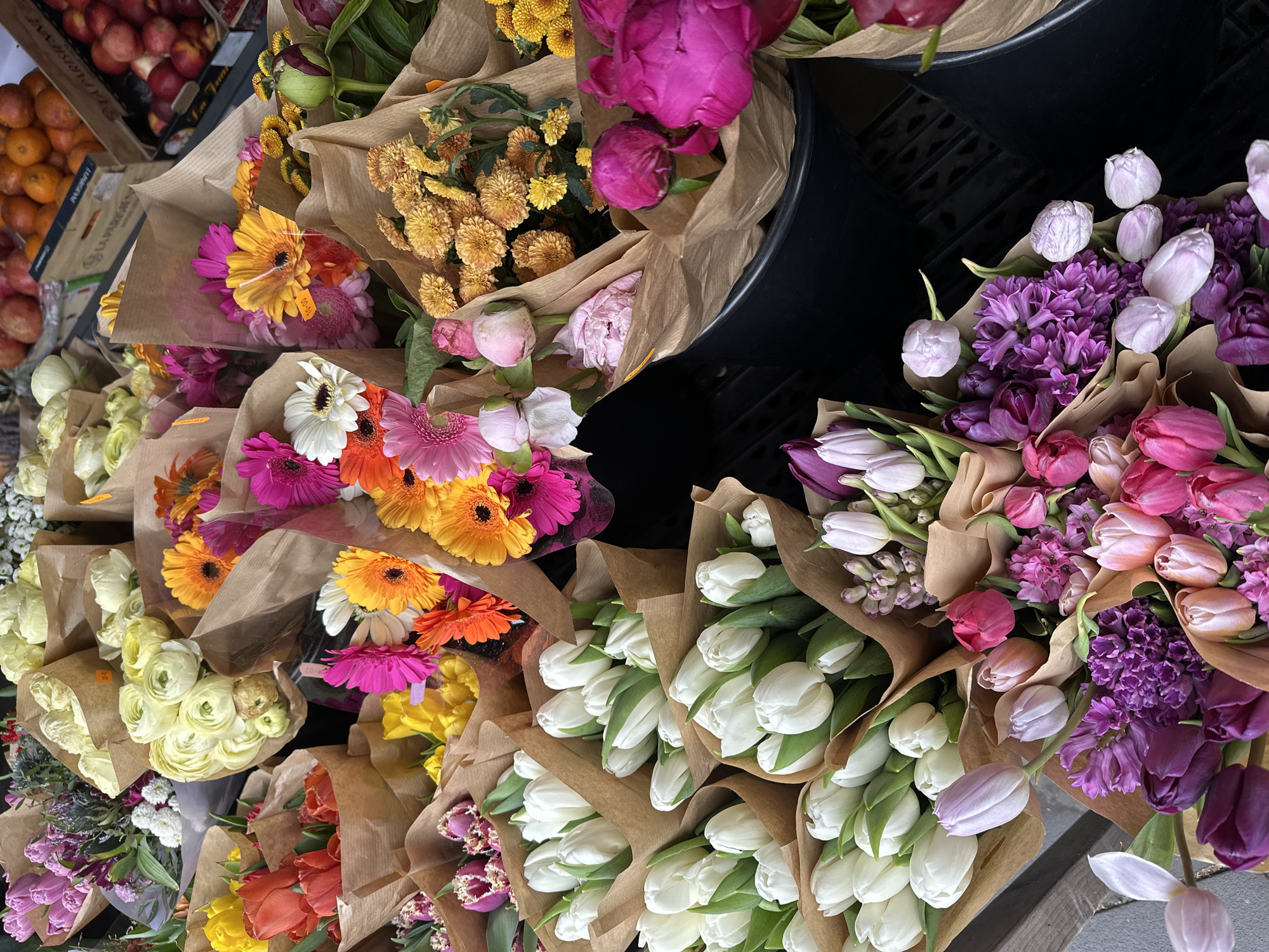 The image captures a flower market scene. Various bouquets of flowers are displayed, including white tulips, pink and purple tulips mixed with hyacinths, orange and pink gerberas, yellow chrysanthemums, and pink peonies, all wrapped in brown paper. Some fruits are visible in the background. The exact location within London is not identifiable from the image. There are no people visible.