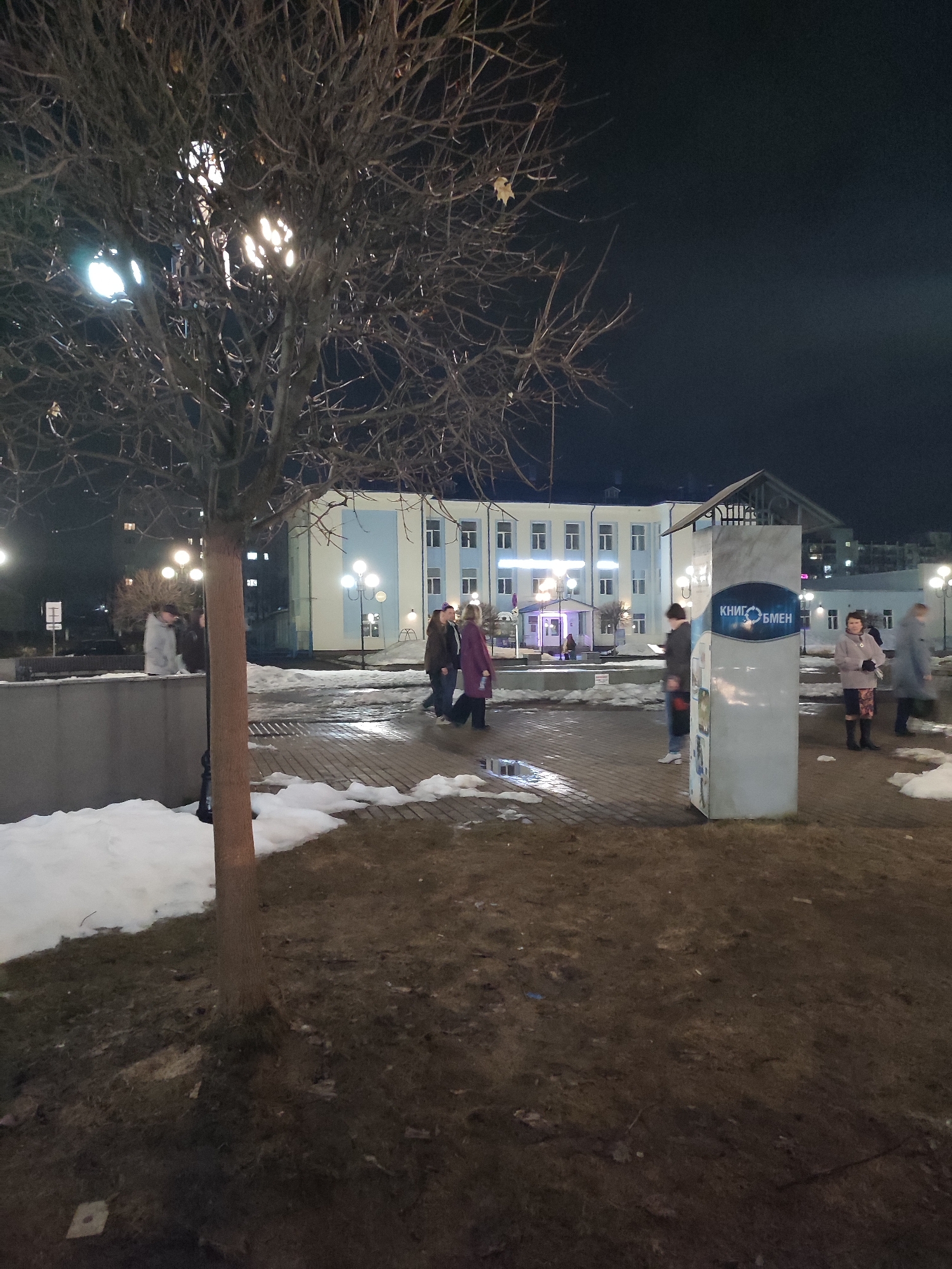 The image shows an outdoor scene at night in what appears to be a public square or park area in Chaikovsky, Russia. A building with light blue and white facade is visible in the background, illuminated by several street lamps. Several people are present in the scene, some walking, others standing. There is a book exchange structure with the Cyrillic text "КНИГОБМЕН" on it. Patches of snow remain on the ground, indicating winter or early spring conditions. A leafless tree stands in the foreground, obscuring the view somewhat. The ground is covered with dirt and dead grass.