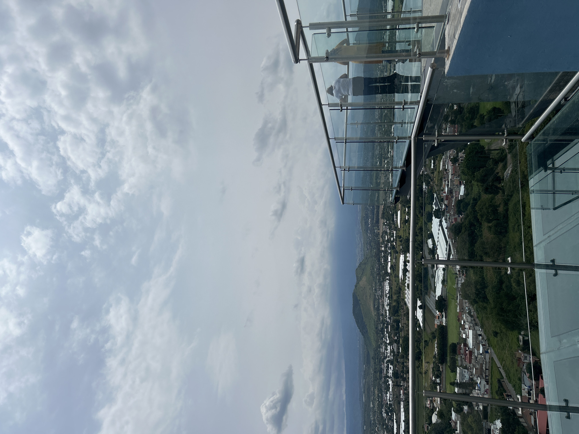 The image shows a panoramic view from a glass-floored observation deck in Atlixco, Mexico. Two individuals are visible from behind, looking out at the landscape. One person is wearing a white shirt and dark pants, while the other is in a light-colored top and pants. The observation deck features modern glass railings with stainless steel accents. Below, the city of Atlixco unfolds, characterized by a mix of residential buildings, green spaces, and agricultural structures, likely greenhouses, with some appearing white. A prominent, rounded green hill is visible in the middle ground, and further in the distance, mountains form the horizon under a cloudy sky. The overall impression is a scenic vista from a high vantage point overlooking the urban and rural expanse of Atlixco.