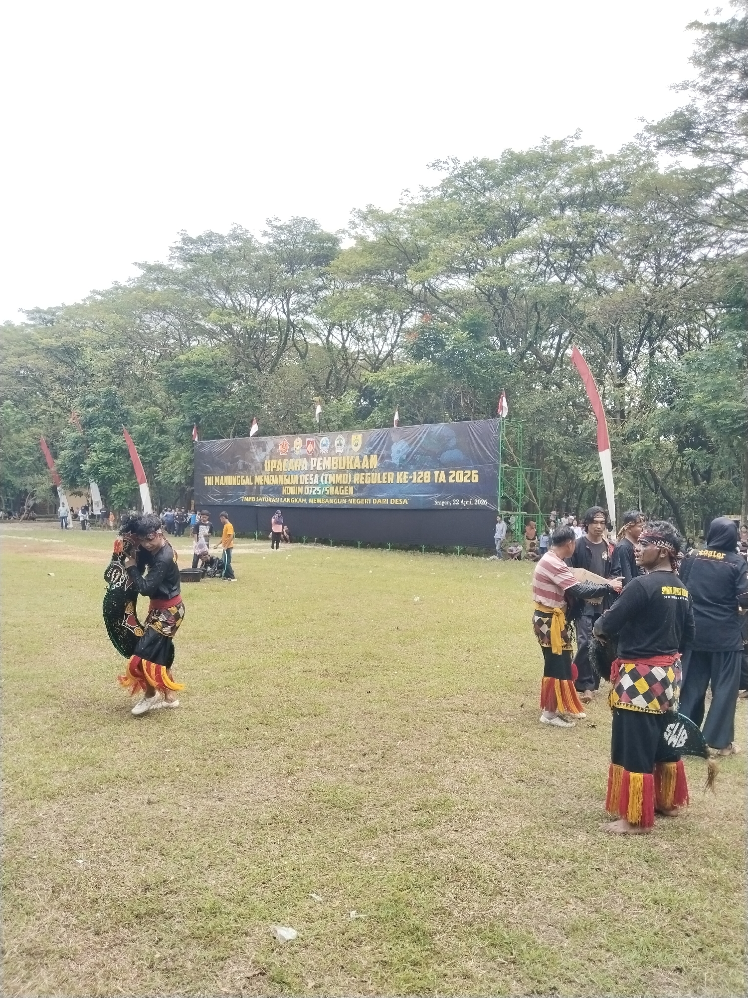 An outdoor opening ceremony for a community development program is taking place on a grassy field in Puro, Sragen, Indonesia, under an overcast sky. Several people are gathered, with some men in traditional attire featuring black tops, patterned sashes, and red and yellow fringed lower garments, appearing to perform or socialize in the foreground. One man is actively engaged in a movement with a decorated object, while others observe or converse.
In the background, a large banner spans the width of the field, displaying the event details. Behind the banner and extending into the background is a dense line of trees, forming a natural backdrop. Red and white flags, resembling the Indonesian national flag colors, are visible on poles around the event area.
The banner prominently announces "UPACARA PEMBUKAAN" (Opening Ceremony) for "TNI MANUNGGAL MEMBANGUN DESA (TMMD) REGULER KE-128 TA 2026" (TNI Manunggal Membangun Desa Regular 128th cycle, Fiscal Year 2026). It also mentions "KODIM 0725/SRAGEN" (Military District Command 0725/Sragen) and the motto "TMMD SATUKAN LANGKAH, MEMBANGUN NEGERI DARI DESA" (TMMD Unites Steps, Builds the Nation from the Village). The event's location and date are specified as "Sragen, 22 April 2026."
The time appears to be daytime, with soft, diffused lighting suggesting an overcast sky. The atmosphere is one of a communal gathering for an official event, possibly involving cultural elements.