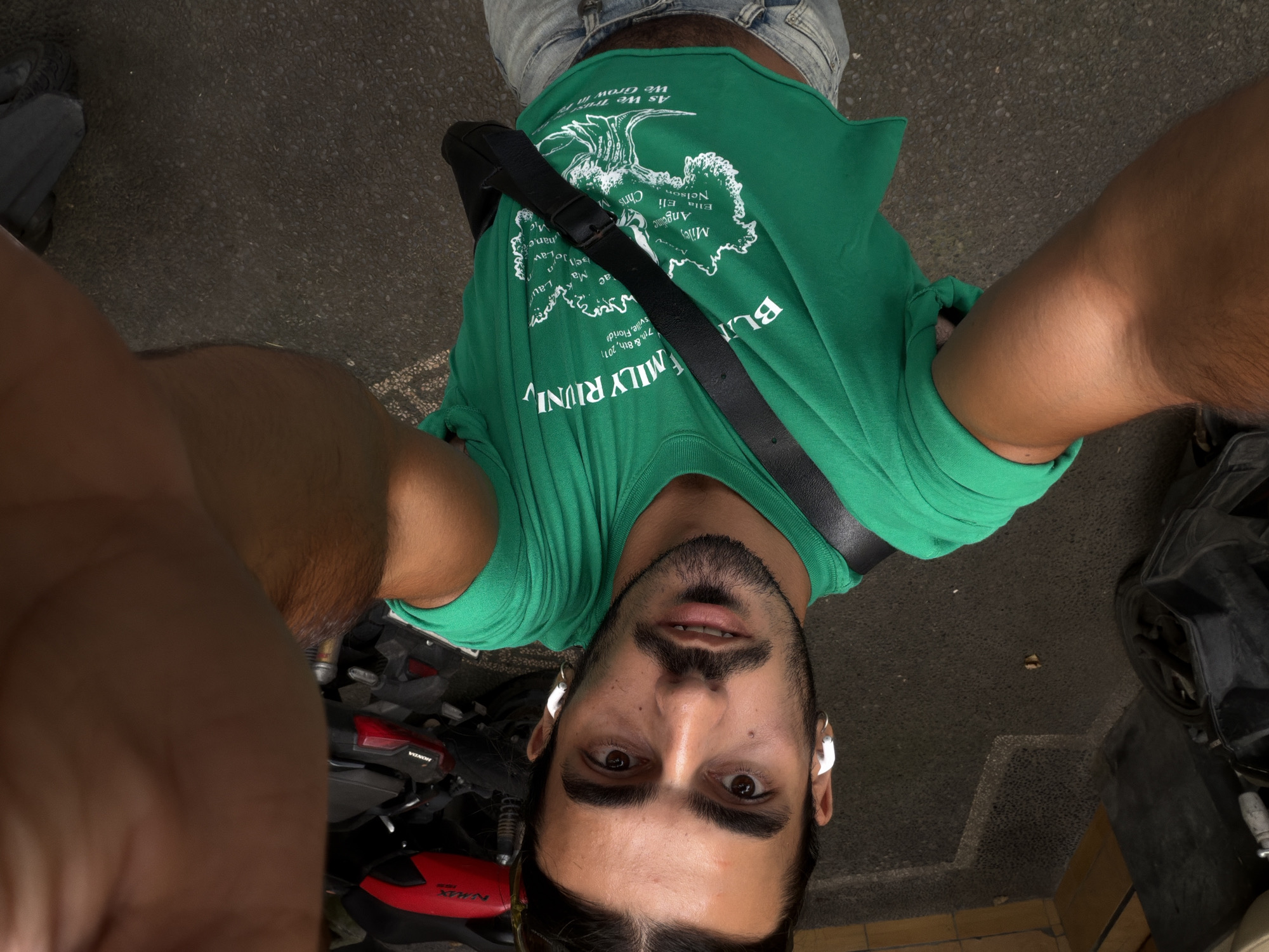 A man with a beard and dark hair wearing a green t-shirt and a black shoulder bag is taking a selfie, likely in an urban area in North Kuta, Indonesia. He is wearing white earbuds. A red and black Honda motorcycle is partially visible in the foreground. A dark object (possibly another vehicle) is partially visible on the right. The ground is paved. The t-shirt has the words "FAMILY RIUNI" and "As We Grow We Trust" written on it.