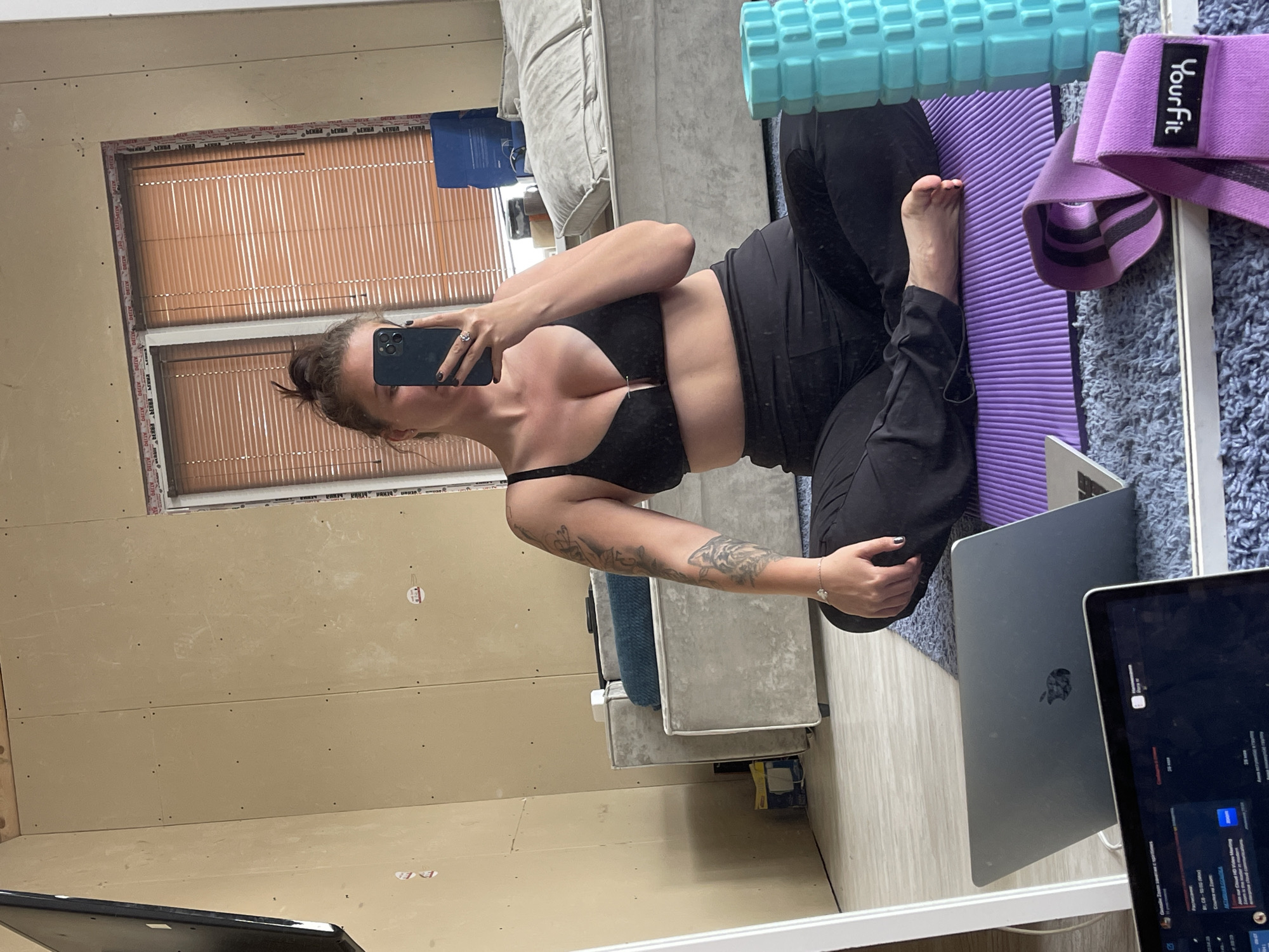 A woman with brown hair in a bun and wearing a black sports bra and black leggings is taking a selfie in a mirror. She is seated on a purple yoga mat with a blue foam roller and a purple resistance band nearby. A laptop is open in front of her. The background appears to be an unfinished room with unpainted drywall walls and a window with blinds. A couch is partially visible in the reflection. The location is unknown.