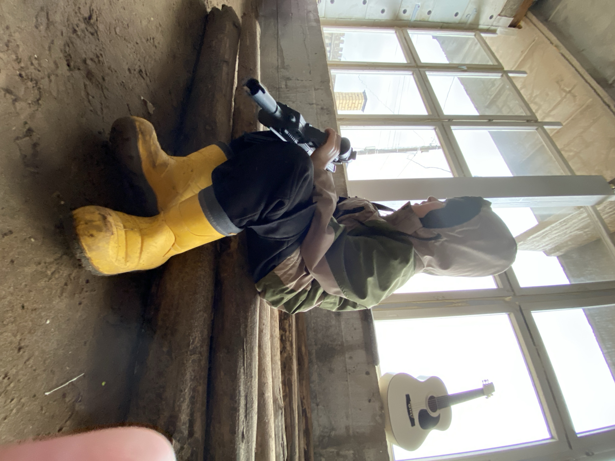 A person wearing yellow boots, dark trousers, a green and beige jacket, and a hoodie, is sitting on a log indoors. They are holding a black object resembling a power tool or weapon. A white guitar is leaning against a wall near a window. The location is Chaikovsky, Russia. The background shows a concrete wall and a window with multiple panes.