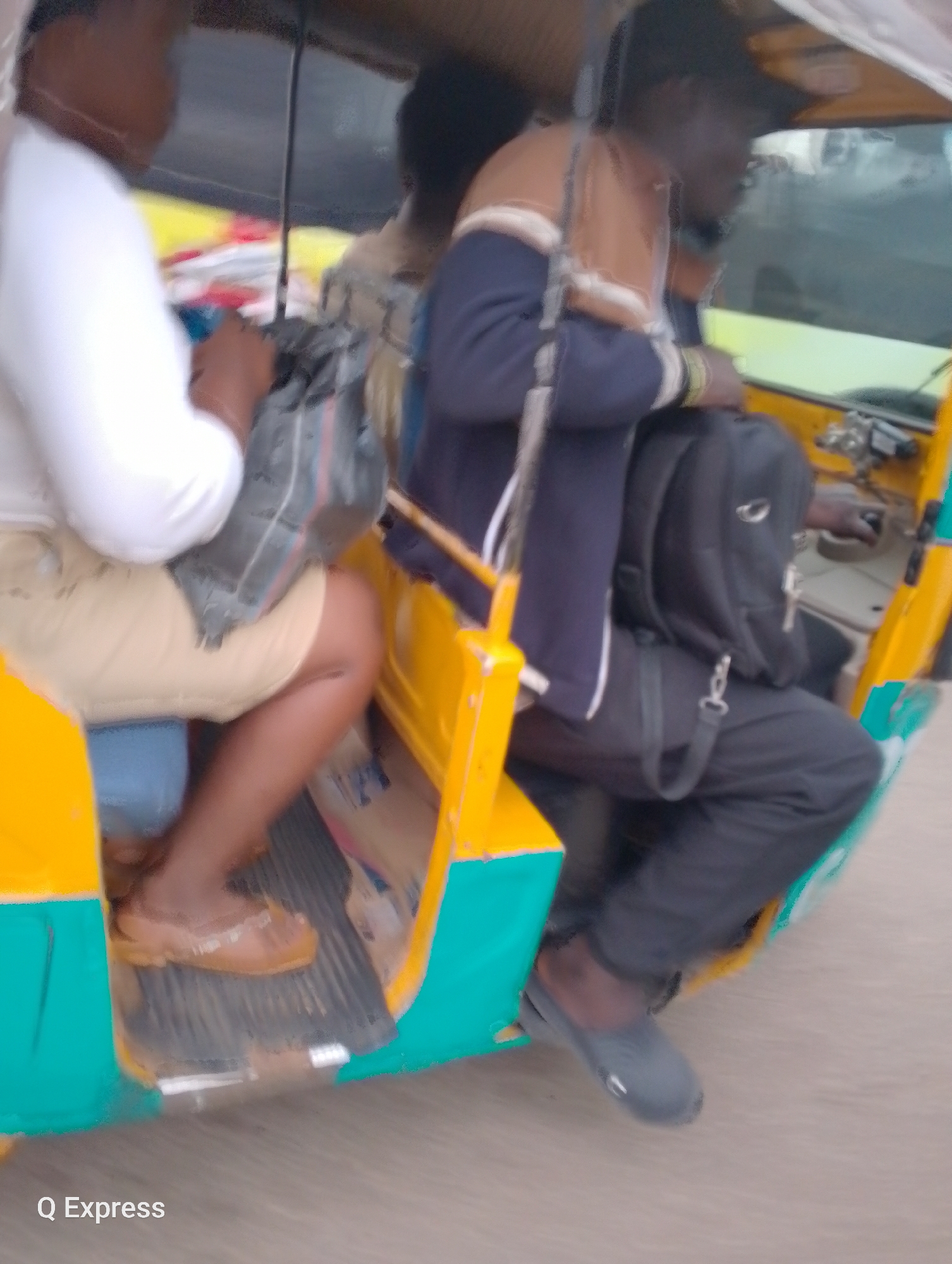 In the city of Jos, Nigeria, a three-wheeled passenger vehicle, commonly known as a "Keke Napep," is captured in motion. The vehicle's exterior is predominantly bright yellow with turquoise accents. Inside, two individuals are visible. The person in the foreground, seated on the left, is wearing a white top and light beige shorts, with a dark bag placed behind them. The driver, on the right, is wearing a navy blue jacket with brown accents and dark trousers, carrying a black backpack. The image appears to be taken from a moving vehicle, indicated by the motion blur throughout the scene. The text "Q Express" is present in the lower left corner, suggesting the name of the transport service. The ground beneath the Keke Napep shows a textured surface.