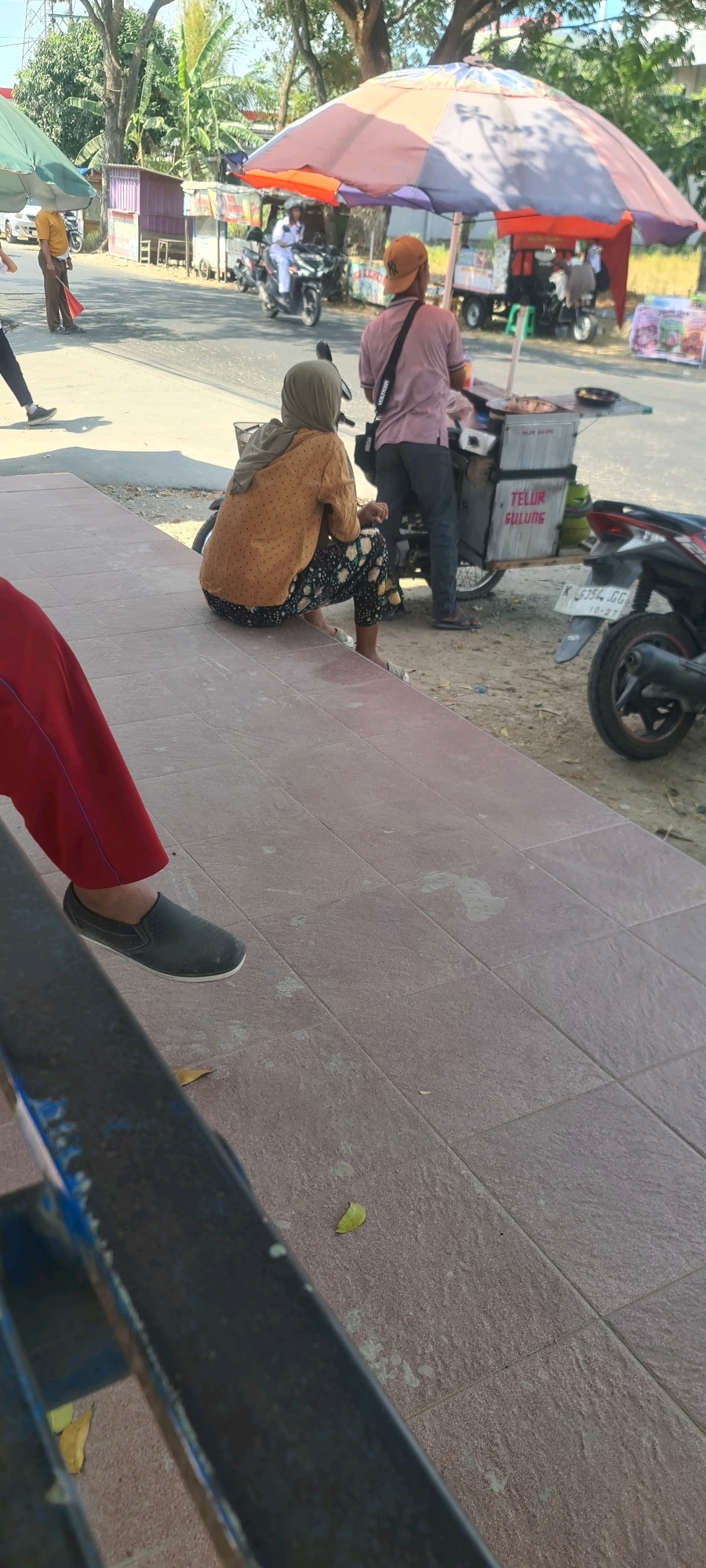 In Jakenan, Indonesia, on a sunny day, an outdoor scene unfolds with activity near a street. A woman, seen from behind, sits on a tiled ledge, wearing a light brown patterned blouse and dark pants with a floral print. She has a light brown hijab covering her hair. Her feet, clad in light-colored slip-on shoes, rest on the ground. Beside her, a dark-colored motorcycle is parked.
In the background, a street vendor operates from a cart under a large, multi-colored umbrella. The cart has signage with the words "TELUR GULUNG". A person wearing a pink shirt and an orange cap stands behind the cart, seemingly attending to customers. Another person in a light-colored uniform rides a motorcycle in the street. Beyond the immediate scene, there are trees, a small building with a purple roof, and other parked vehicles. The ground is a mix of pavement and dirt. The overall impression is of a typical roadside scene with local commerce.