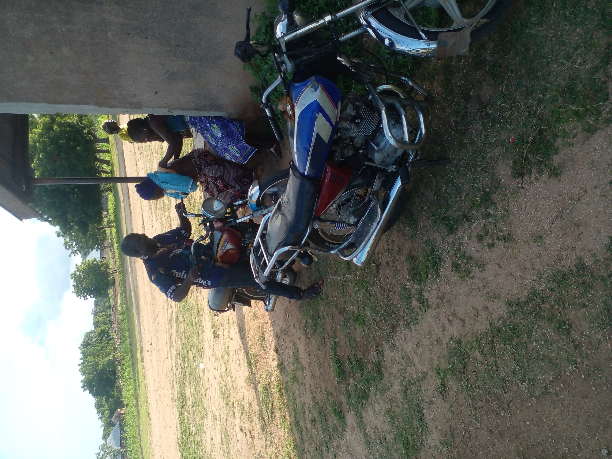 In an outdoor scene, a man in a patterned shirt and dark pants stands next to a motorcycle. To his right, three women are seated under a covered structure. One woman, wearing a blue headscarf and patterned skirt, is interacting with another woman in a blue top and patterned skirt. A third woman in a yellow top is also present. Two motorcycles are visible, one in the foreground with blue and white accents on its fuel tank and another behind it with red and maroon colors. The environment is dusty with sparse green grass and trees in the background, suggesting a rural or semi-urban setting. The lighting indicates it is daytime, likely late afternoon or early morning.