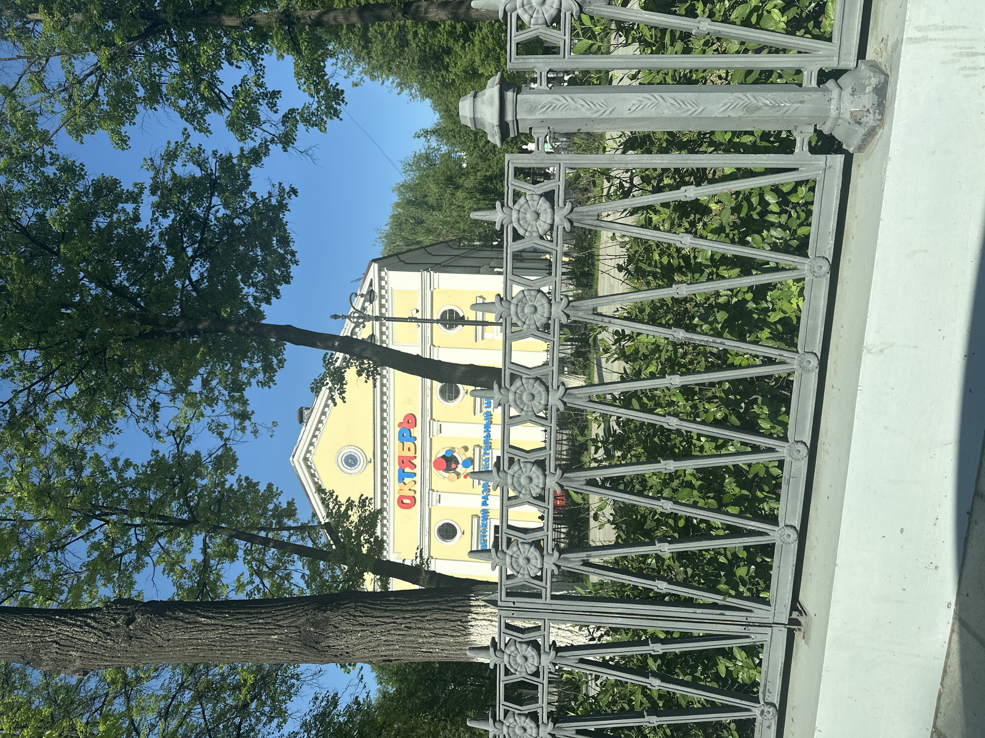 The image shows the Perm Children's Railway building, located in the city of Perm, Russia, as viewed through a metal fence. The building is painted yellow and has the word "Октябрь" (October) written in red letters above an image of stylized colorful atoms. The surrounding environment includes trees and greenery. The image is taken during the daytime under a blue sky.
