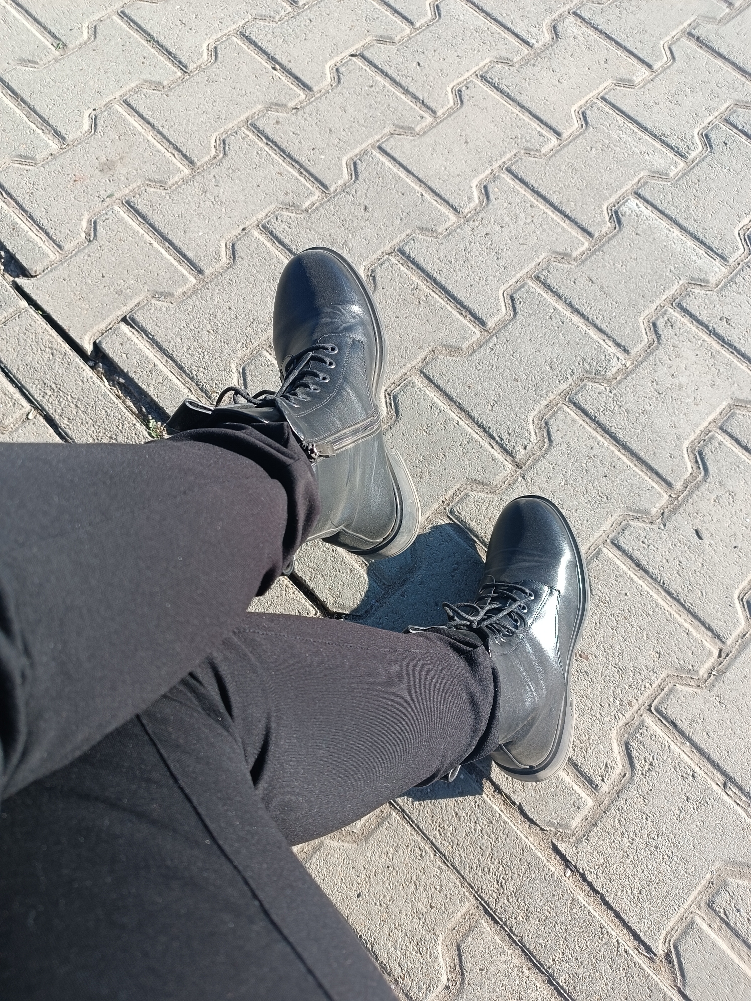 The image shows a first-person perspective looking down at a person's legs and feet. The subject is wearing black pants and black combat-style boots. The legs are extended, resting on a light grey paved surface.
The setting is outdoors, likely a sidewalk or pedestrian area in Chaikovsky, Russia. The ground is covered with interlocking concrete pavers arranged in a distinct herringbone or zigzag pattern. The texture of the pavers is coarse and rugged.
The pants are dark, appearing to be made of a slightly textured fabric. The boots are black, with visible laces and eyelets, and feature a zipper along the inner ankle, suggesting ease of wear. They have a subtle sheen, reflecting the bright light.
The scene is illuminated by strong, clear sunlight, casting sharp shadows from the boots and legs onto the pavement. This indicates a bright, sunny daytime. There is no discernible activity beyond the person resting or pausing. No text is visible in the image. The overall impression is a casual, everyday moment captured in an urban environment.