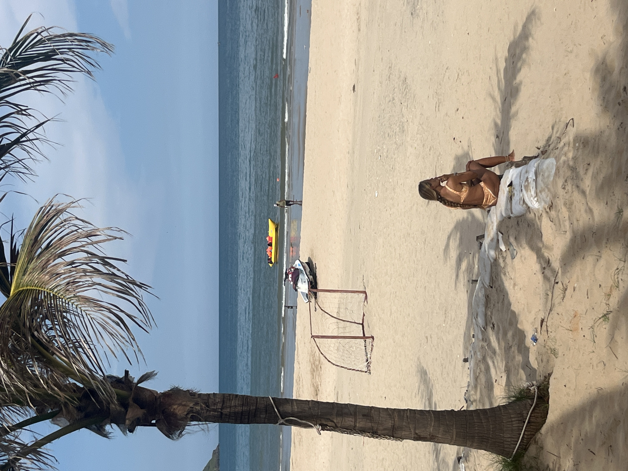 In Mui Ne, Vietnam, a woman with blonde hair braided down her back sits on sandbags on a beach. She is wearing a yellow bikini and looking out at the ocean. In the foreground, a palm tree's shadow falls across the sand. The ocean is calm, and a yellow inflatable boat with several people wearing orange life jackets is visible on the water. A white jet ski is parked on the sand near a small soccer net. The sky is blue with a few scattered clouds.