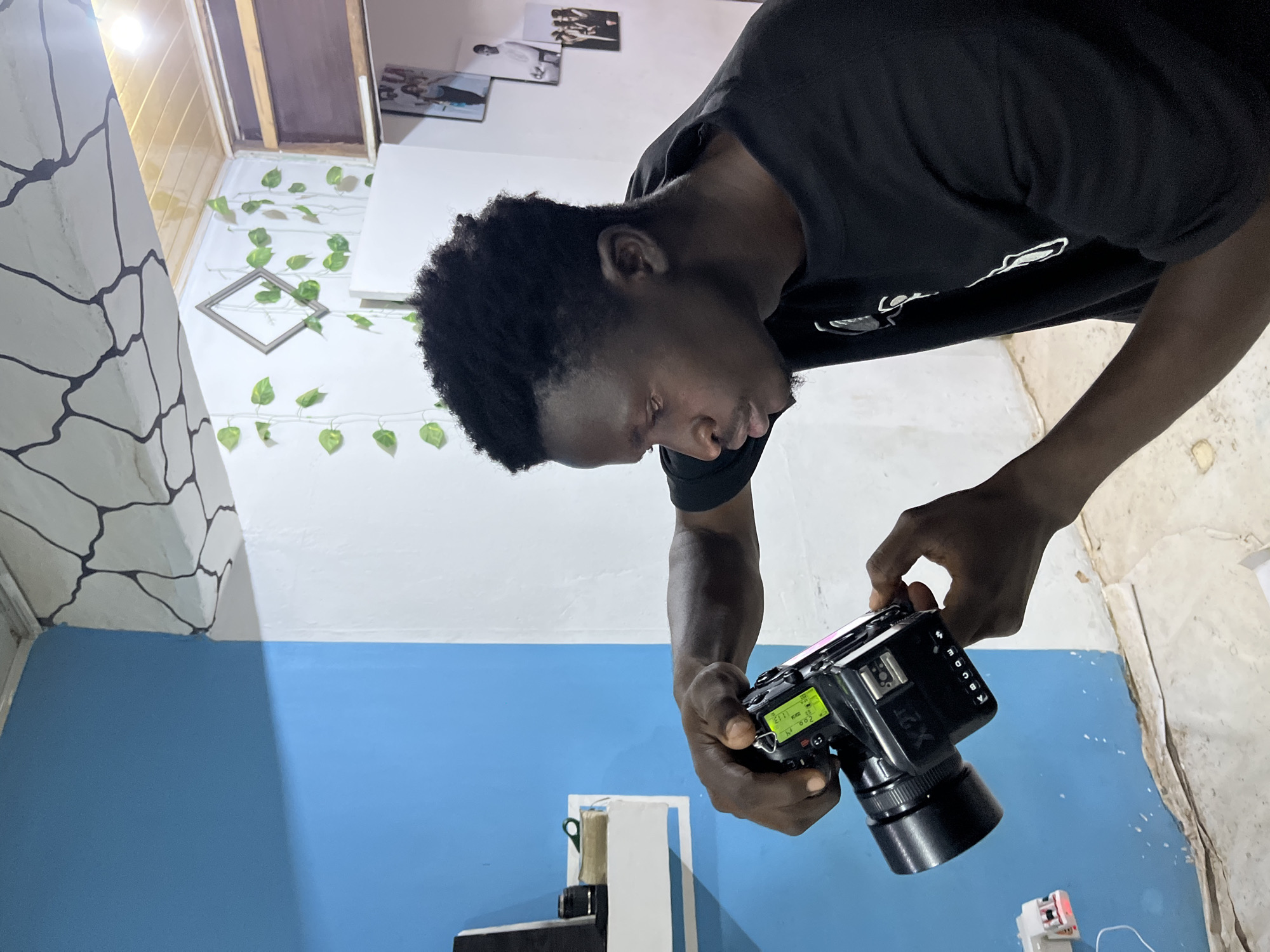 A man with dark hair and a black T-shirt is holding a professional camera. He is looking down at the camera screen, adjusting settings. The background features a wall divided into blue and white sections, with a decorative vine and a framed picture. There are other pictures hanging on the wall. A stone pattern is visible on one side of the wall. The location is Gura Top, Nigeria.
