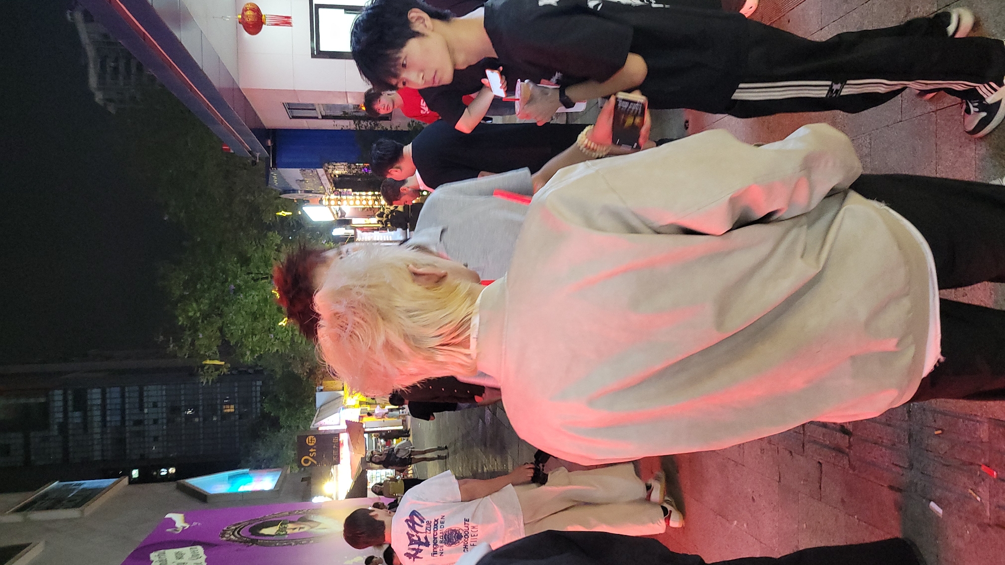 Here is a concise description of the image:
A street scene at night in what appears to be a city in Asia. Several people are standing and using their phones. One person has bright blonde hair and is wearing a light-colored jacket. Another person is wearing black pants with white stripes. There are buildings and streetlights visible in the background. There is a red lantern hung near a building.