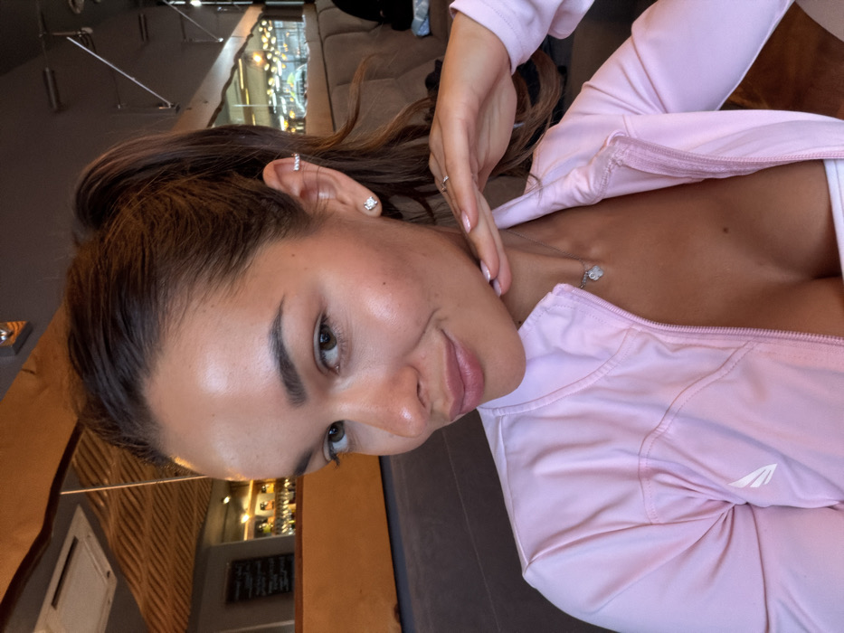 A female individual, appearing to be in her early to mid-20s, is captured in a medium close-up, indoors. She has tan skin, dark brown hair pulled into a high ponytail, and brown eyes. She exhibits a faint smile while looking directly forward. Her left hand, with manicured nails painted in a light hue and a silver ring on the ring finger, is positioned with fingertips touching her jawline/chin. She is wearing a light pink athletic jacket, partially unzipped, revealing a white undergarment. A small white winged logo is present on the left chest area of the jacket. A silver necklace with a light-colored, possibly heart-shaped, pendant is visible. Her right ear features a square-cut clear stud earring in the lobe and two thin silver cuff earrings on the helix.
The background is blurred, indicating an indoor commercial establishment, likely a cafe or restaurant. To the upper left, a reflective surface (possibly a mirror or polished panel) shows a reflection of the ceiling and overhead lights. Below this, a dark-colored menu board with white writing is partially visible. Behind the subject and extending to the right, a long segment of cushioned booth seating, upholstered in a brown or tan fabric, is present. Further in the background, indistinct shapes of bottles and glasses suggest a bar area. The lighting is ambient and consistent with an indoor setting.
The specific location within the city of Krasnoyarsk, Russia, cannot be determined from the visual information available in the image.