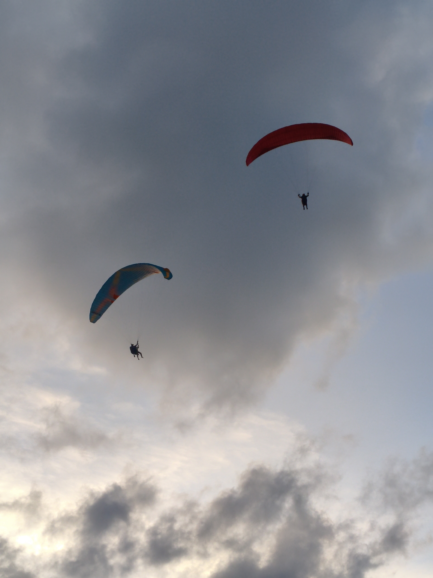 Two paragliders are visible against a cloudy sky, over Rawai, Thailand. The paraglider on the left has a blue and orange wing and is lower in the frame. The paraglider on the right has a red wing and is higher in the frame. The sky is a mix of dark and light clouds, suggesting either dawn or dusk.