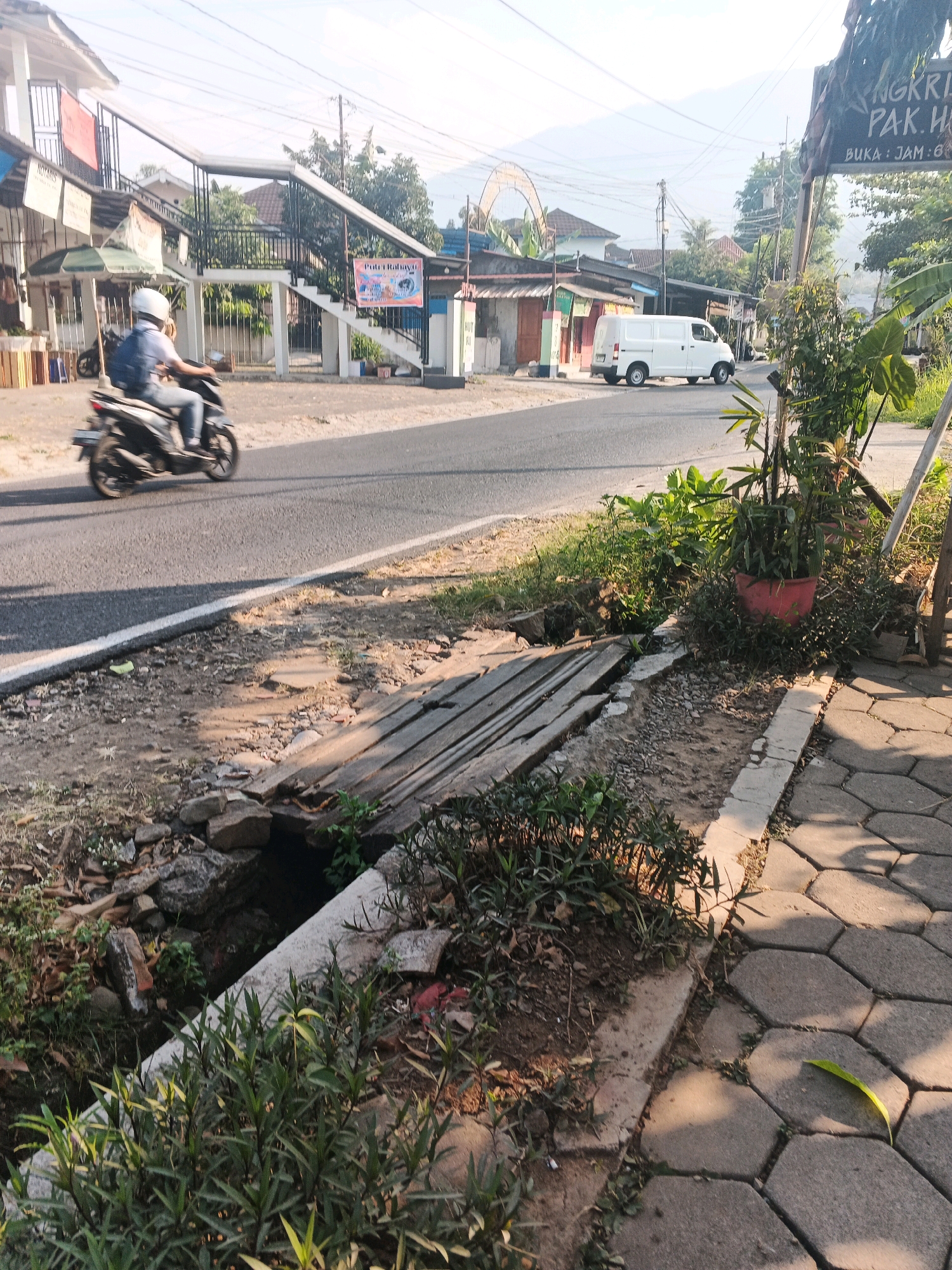 This image depicts a street scene in Ungaran, Indonesia. In the foreground, a small, unpaved ditch runs along the side of a paved walkway. The ditch contains some debris, rocks, and sparse vegetation. A makeshift bridge, constructed from wooden planks, crosses the ditch. Beyond the walkway and ditch, a paved road is visible, with a person riding a motorcycle towards the left side of the frame. The rider is wearing a white helmet and a blue backpack. On the other side of the road, there are several buildings, including a white building with a pedestrian overpass leading to it. A white van is driving on the road, moving away from the viewer towards the right side of the frame. Overhead power lines are present in the scene. Lush green foliage, including plants in a red pot, is situated to the right of the walkway. A sign with Indonesian text is also visible on the right. The overall impression is a moderately developed urban or semi-urban area.