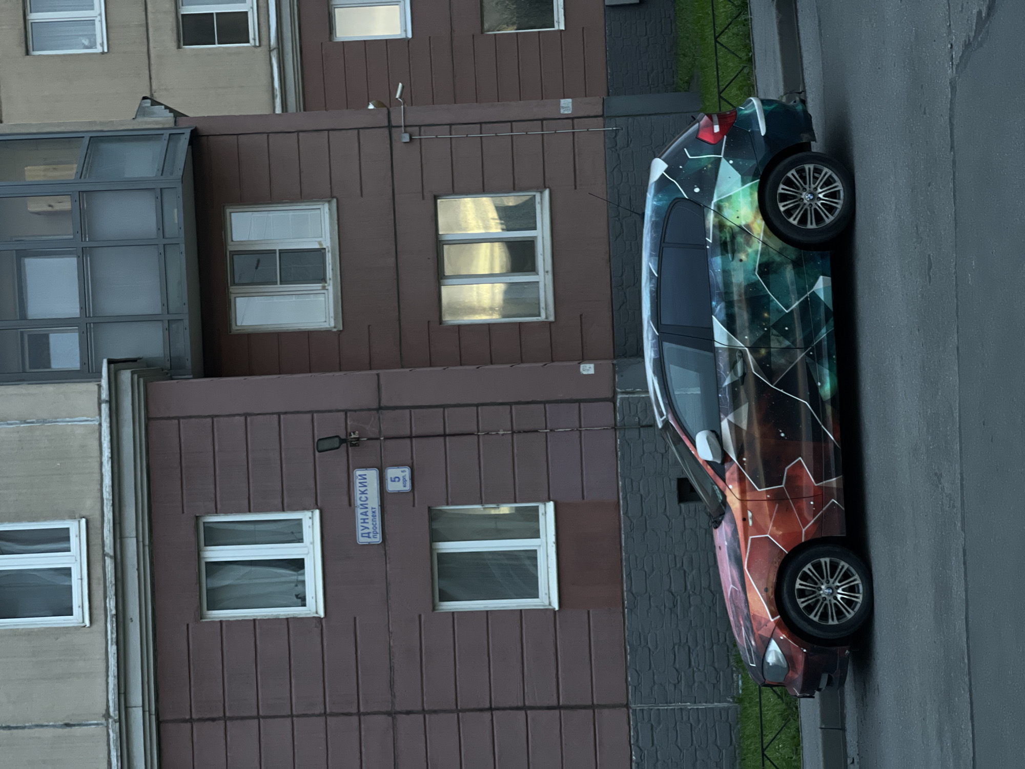 The image shows a side view of a hatchback car parked on a street. The car has a custom wrap with a colorful, geometric, cosmic-themed design. The car is parked in front of a multi-story apartment building. The building has brown and grey facades, with several windows visible. A street sign on the building reads "ДУНАЙСКИЙ проспект" (Dunaysky Prospekt) and "5 корп. 5" (Building 5, Corp. 5). A black metal fence separates the sidewalk from a grassy area behind the car. The sky is overcast, with reflections of sunlight visible in some of the windows. The overall scene depicts an urban environment.