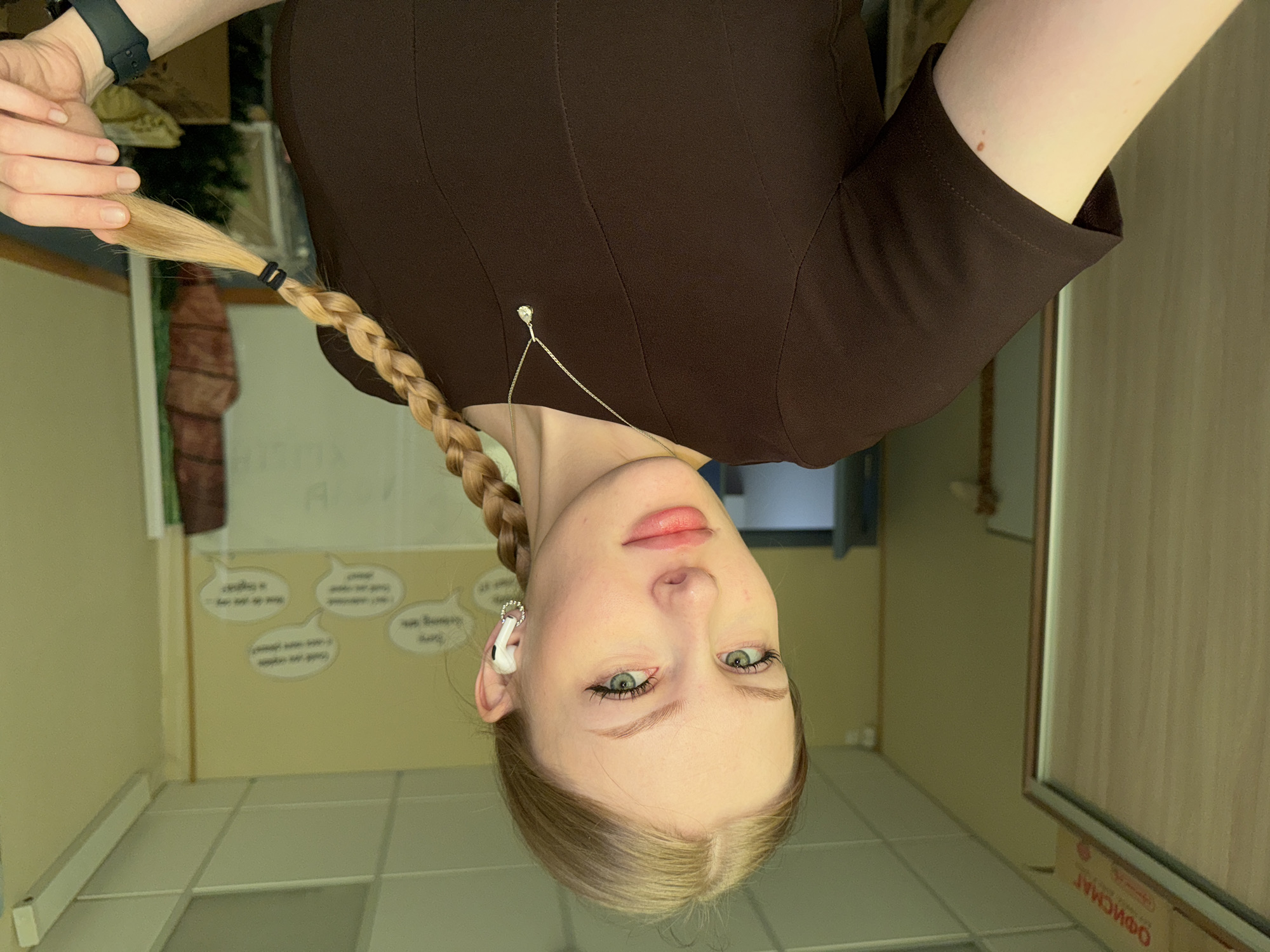A young, light-skinned female with blonde hair braided and falling towards the camera is facing downward. She has green eyes and is wearing a brown short-sleeved dress and a silver necklace. There appears to be an earbud in her right ear. The background shows what appears to be a room with tile floors and walls. A portion of a window and a wall with word bubbles are visible. The location is in Perm, Russia.