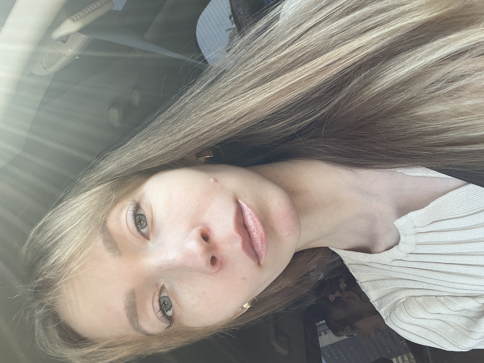A young woman with long, blonde hair is taking a selfie inside a vehicle, possibly a car. The photo appears to be taken during daylight hours, based on the lighting. The interior of the vehicle is visible in the background, including the rearview mirror and other dashboard elements. The woman is wearing a light-colored, ribbed top. She has fair skin, light-colored eyes, and is wearing light makeup, including lipstick. Based on the reflection in the surface behind her, there is evidence of an urban environment, potentially city streets. No explicit details confirm the precise location is Perm, Russia.