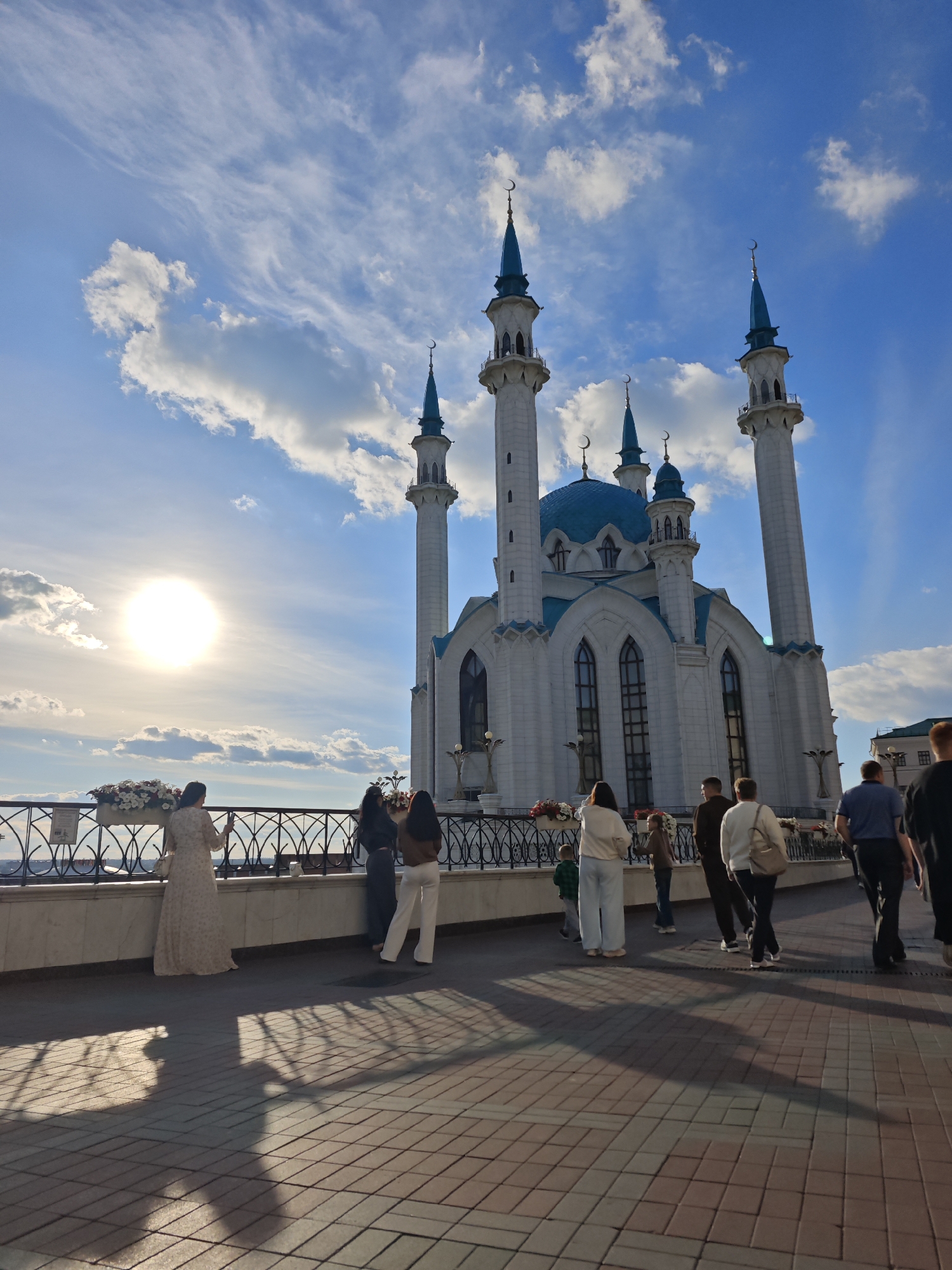 This image captures the Kul Sharif Mosque in Kazan, Russia, on a bright, sunny day with a partly cloudy sky. The sun is prominently visible on the left side of the frame, casting long shadows on the paved walkway in the foreground. Several people are present, appearing to be tourists or visitors, as they stand near a decorative railing and observe the mosque.
The mosque itself is a magnificent structure with white walls, light blue domes, and tall minarets topped with crescent moons. Its intricate architectural details are clearly visible, highlighted by the sunlight.
The people in the image are casually dressed, and their postures suggest they are enjoying the view and possibly taking photographs. A woman in a long, light-colored dress stands out as she holds up her phone, likely capturing a moment. Other individuals are seen walking or standing in groups, some facing the mosque, others in profile. There is a sense of calm and appreciation for the beautiful scenery. The time of day appears to be late afternoon or early evening, given the angle of the sun and the length of the shadows. The weather is pleasant, with a gentle breeze suggested by the wispy clouds in the sky. No visible text is present in the image.