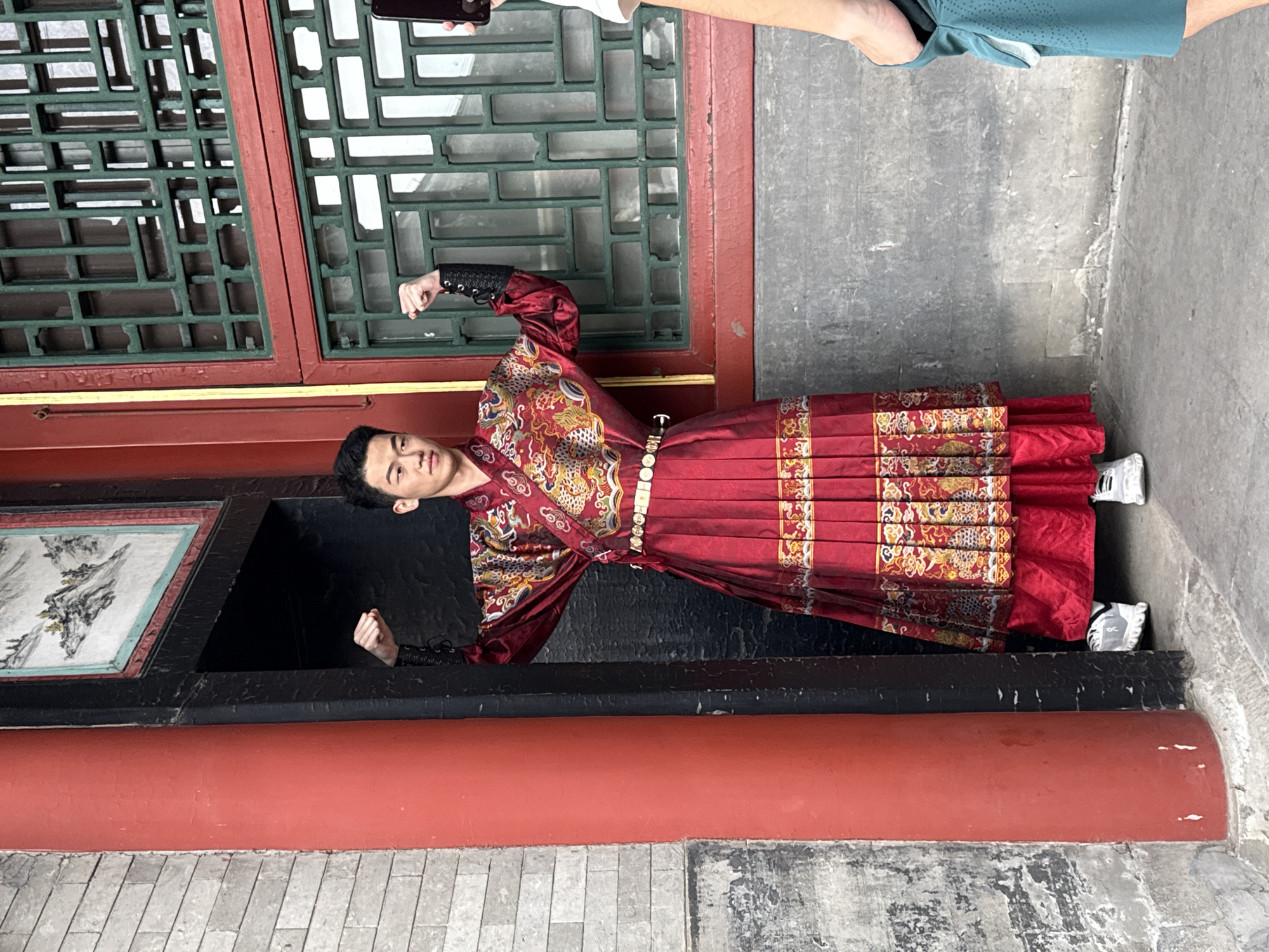 The image displays a young East Asian male posing in a doorway within a traditional Chinese architectural setting in Haidian District, China. The man, appearing in his late teens or early twenties, wears an elaborate, dark red traditional garment, possibly a Hanfu, richly embroidered with gold and colorful dragon and auspicious patterns. His attire includes a pleated lower skirt-like section and an upper robe with wide sleeves, cinched by a decorative white-buckled belt. Modern white and grey athletic sneakers are visible on his feet. He stands with both arms bent at the elbows, fists clenched, and a focused expression directed slightly left of the viewer.
To the left of the doorway, a smooth, cylindrical red pillar stands against an aged grey brick wall. Above the doorway, a framed traditional Chinese landscape painting, featuring mountains and water, is integrated into the wall. To the right of the doorway, a wall section contains two stacked windows, each fitted with intricate dark green wooden lattice patterns set within red frames. On the far right, the right arm and shoulder of a second person are partially visible. This individual holds a black smartphone, aimed at the posing man, indicating they are taking a photograph. The photographer is wearing a light-colored top and a teal athletic short or skirt. The ground is a light grey concrete surface, showing signs of wear.
