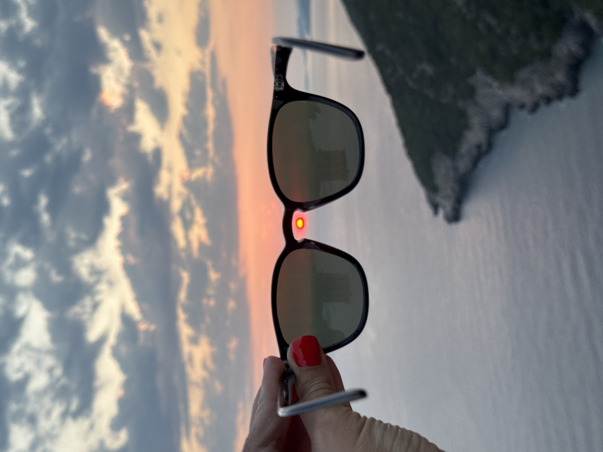A person's hand with bright red nail polish is holding up a pair of black-framed sunglasses. The sunglasses are positioned in front of a scenic sunset over a body of water, possibly the Adriatic Sea, with a rugged, tree-covered coastline visible on the right. The sun, a vibrant red orb, is framed perfectly within the right lens of the sunglasses, creating a dramatic effect. The sky above is filled with a mix of soft orange, pink, and gray clouds, indicative of a sunset. The water below is calm and reflects the colors of the sky. The general location is likely near the city of Zagora, Montenegro, given the coastal landscape and typical sunset views in that region.