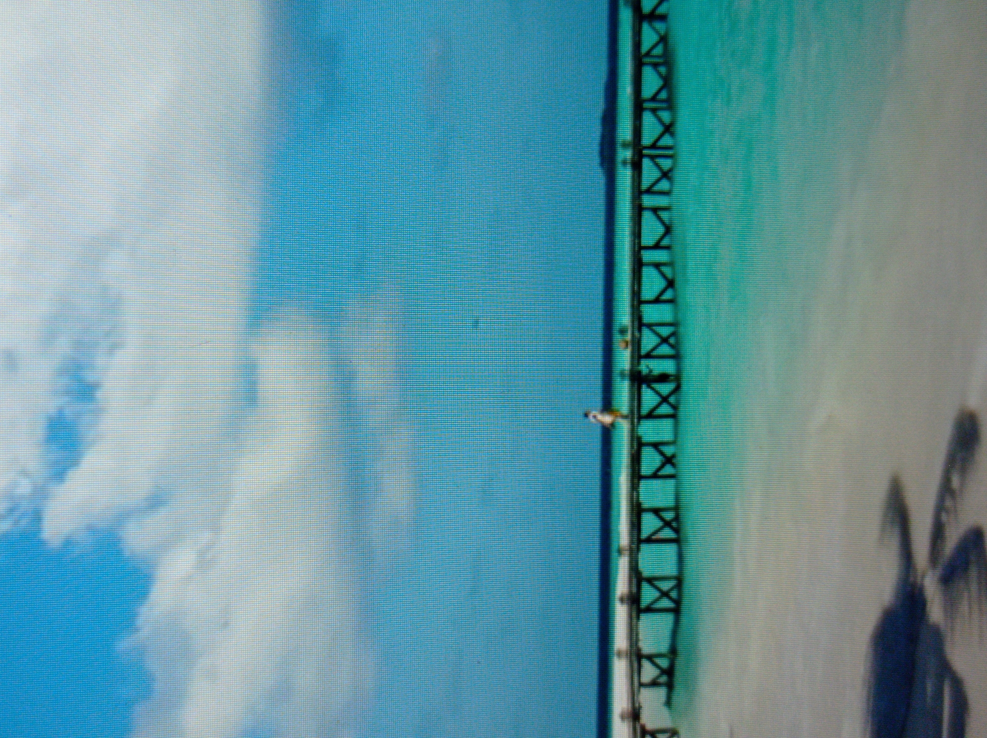 A wooden pier extends over a turquoise body of water under a bright blue sky with scattered white clouds. A lone person, dressed in light clothing, walks on the pier. The foreground shows a sandy beach with a shadow cast from an unseen object. The image appears to be a digital display due to visible pixelation. The scene does not depict any location in Mukachevo, Ukraine.