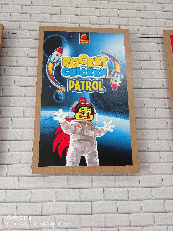 A poster for "Rocket Chicken Patrol" is mounted on a white brick wall. The poster features a cartoon chicken wearing a spacesuit and a red cape, standing in front of a blue space background with three red, white, and blue rockets. The poster is mounted on a wooden frame. The poster is located in Patrol, Indonesia, at the coordinates -6.318 latitude and 107.986 longitude. The poster is the subject of the image.