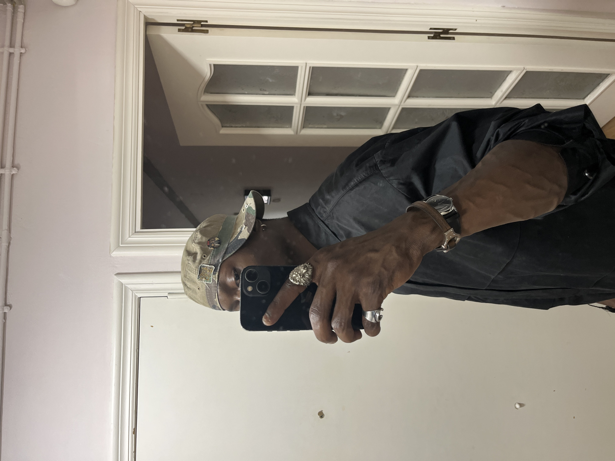 This media file captures a mirror selfie taken by an individual in an indoor setting, identified contextually as Barcelona, Spain. The person is visible from the chest up in the reflection, holding a black smartphone (likely an iPhone) with their right hand to capture the image.
The individual is wearing a black collared shirt and a camouflage-patterned baseball cap adorned with several small pins or patches. Accessories include a wristwatch with a brown leather strap on their left wrist, and two prominent silver rings on their right hand: one large and intricately designed (possibly depicting a lion's head), and another simpler, silver band. The person's face is mostly obscured by the phone, with only part of the right eye and forehead visible above it.
The mirror is framed by white molding. Reflected behind the person is the upper section of a white door, distinguished by multiple rectangular glass panes and a gold-colored hinge near the top. To the left of the mirror, the wall outside the reflection is a light pink or lavender hue, with white vertical radiator pipes running along it. The lighting suggests an indoor environment, but specific time of day or weather cannot be determined. No explicit text is legible within the image.