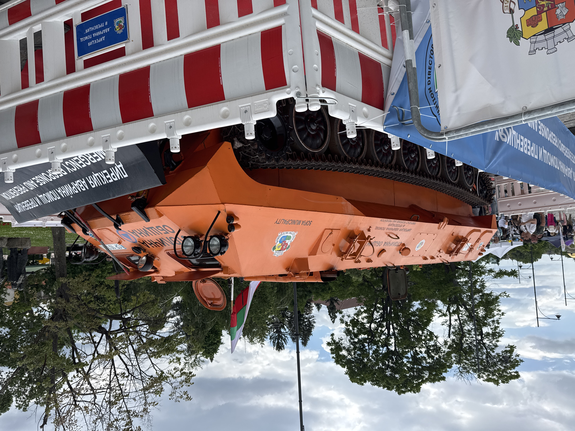 The image shows an orange vehicle with tracks, possibly an emergency response vehicle, as indicated by the text "EMERGENCY RESPONSE AND PREVENTION" and "ДИРЕКЦИЯ АВАРИЙНА ПОМОЩ И ПРЕВЕНЦИЯ". The vehicle has "SOFIA MUNICIPALITY" printed on its side. It is surrounded by barriers with red and white stripes. A Bulgarian flag is visible. Trees and a cloudy sky are in the background. There are flags and banners with text on them. People can be seen standing in the background.
