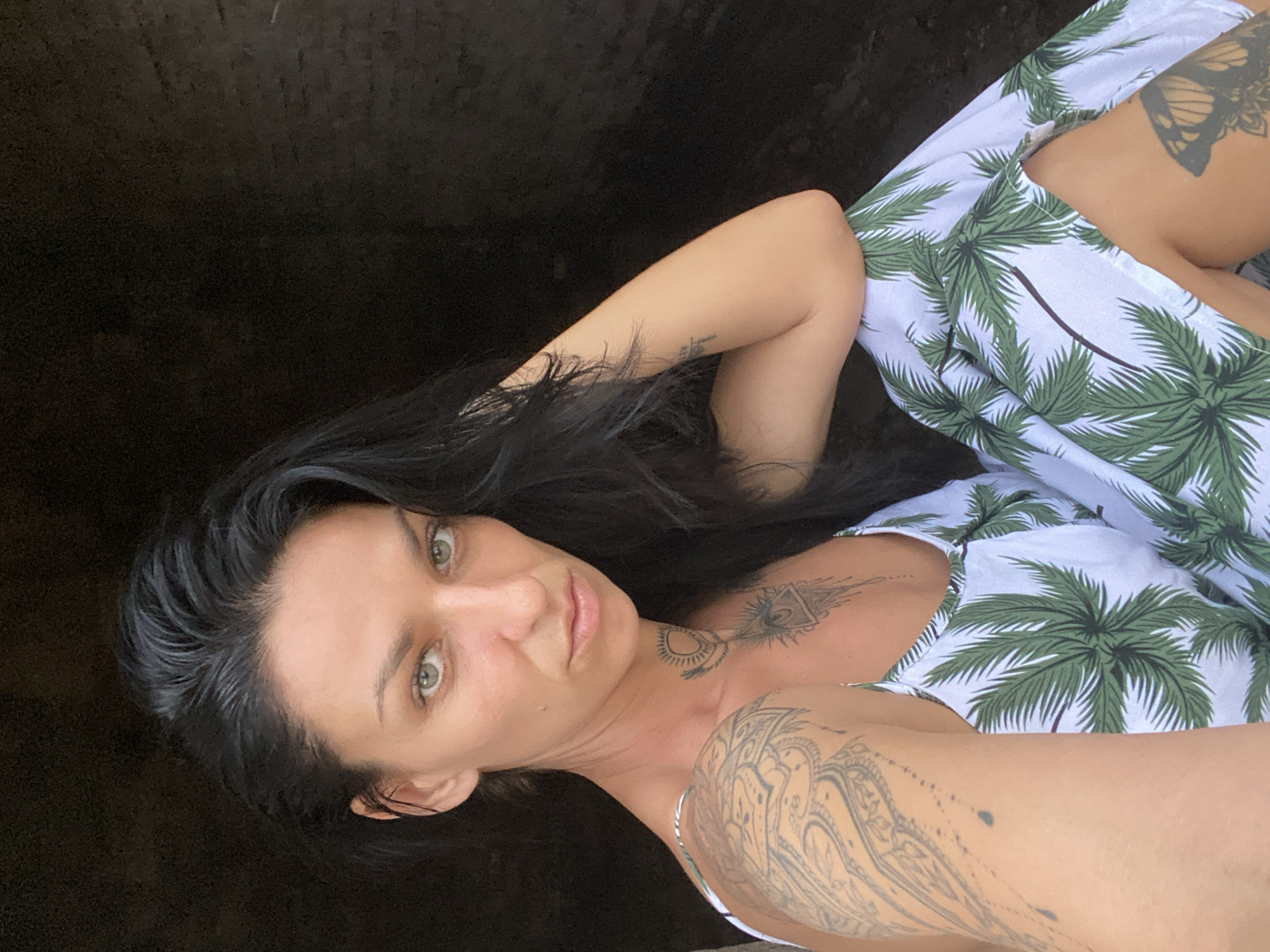 A woman with dark hair and light eyes is lying down, facing the camera, in what appears to be Phu Thuy Ward, Vietnam. She is wearing a white dress with a green palm tree pattern. The background is dark and blurry. The woman has tattoos on her arms, chest, and thigh. She is holding her arm up near her head.