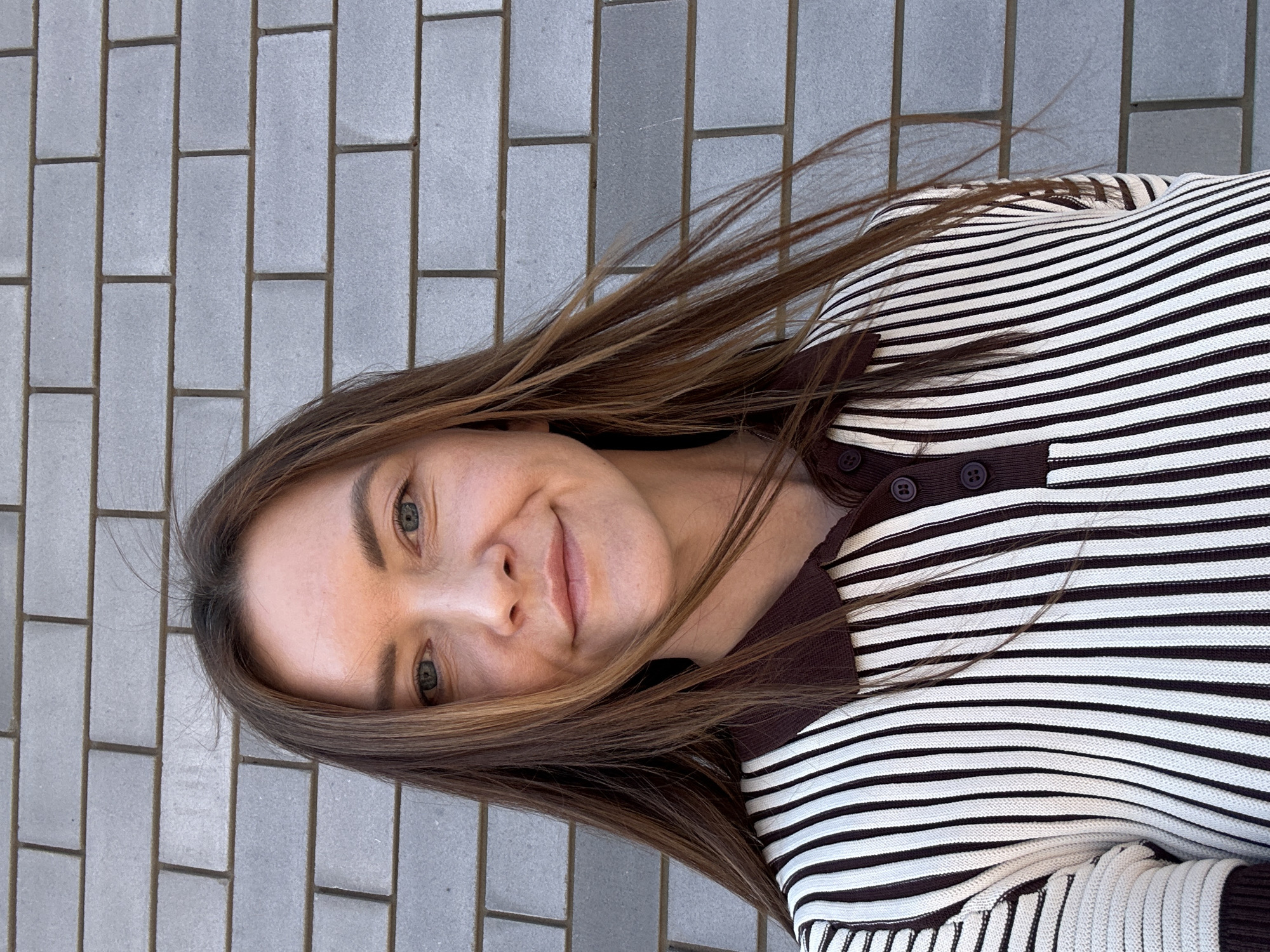 A woman is outdoors, lying on a gray brick sidewalk. Her head is tilted to the left. She has brown hair and is wearing a brown and white striped shirt with buttons at the collar. The location is likely Прикамский, Russia.
