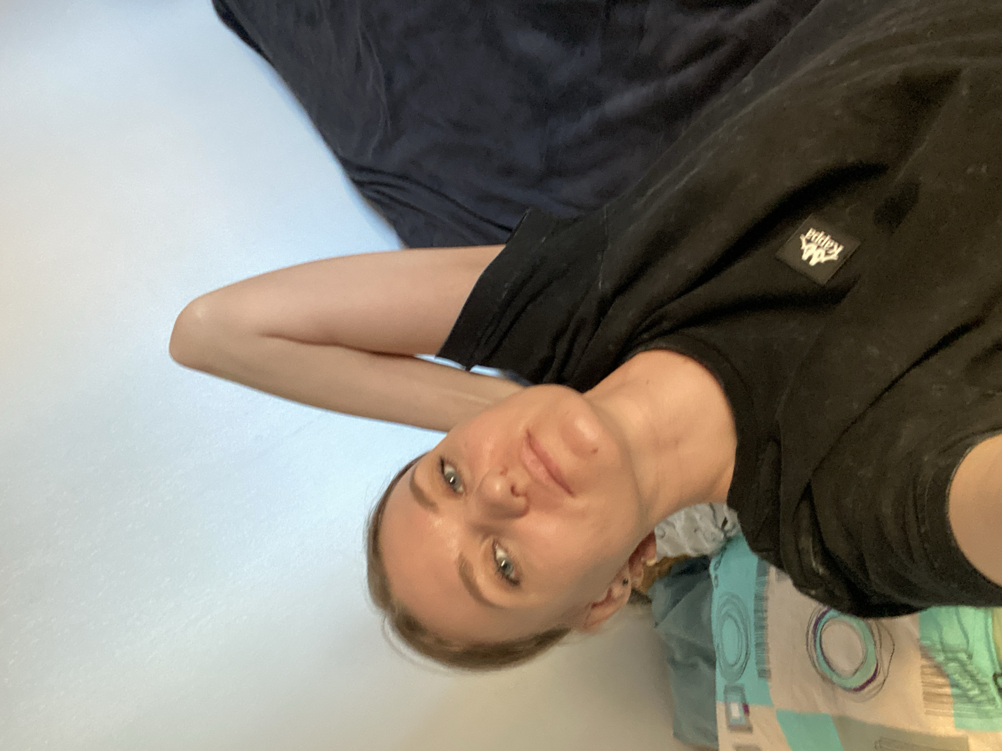 A selfie of a Caucasian female with fair skin, light brown hair pulled back, and blue eyes, wearing a black t-shirt with a Kappa logo. She is holding one arm behind her head. She is laying on bedding consisting of a dark gray blanket and patterned sheets. The setting appears to be indoors against a white wall. There is no evidence to indicate this image was taken in Saint Petersburg, Russia.