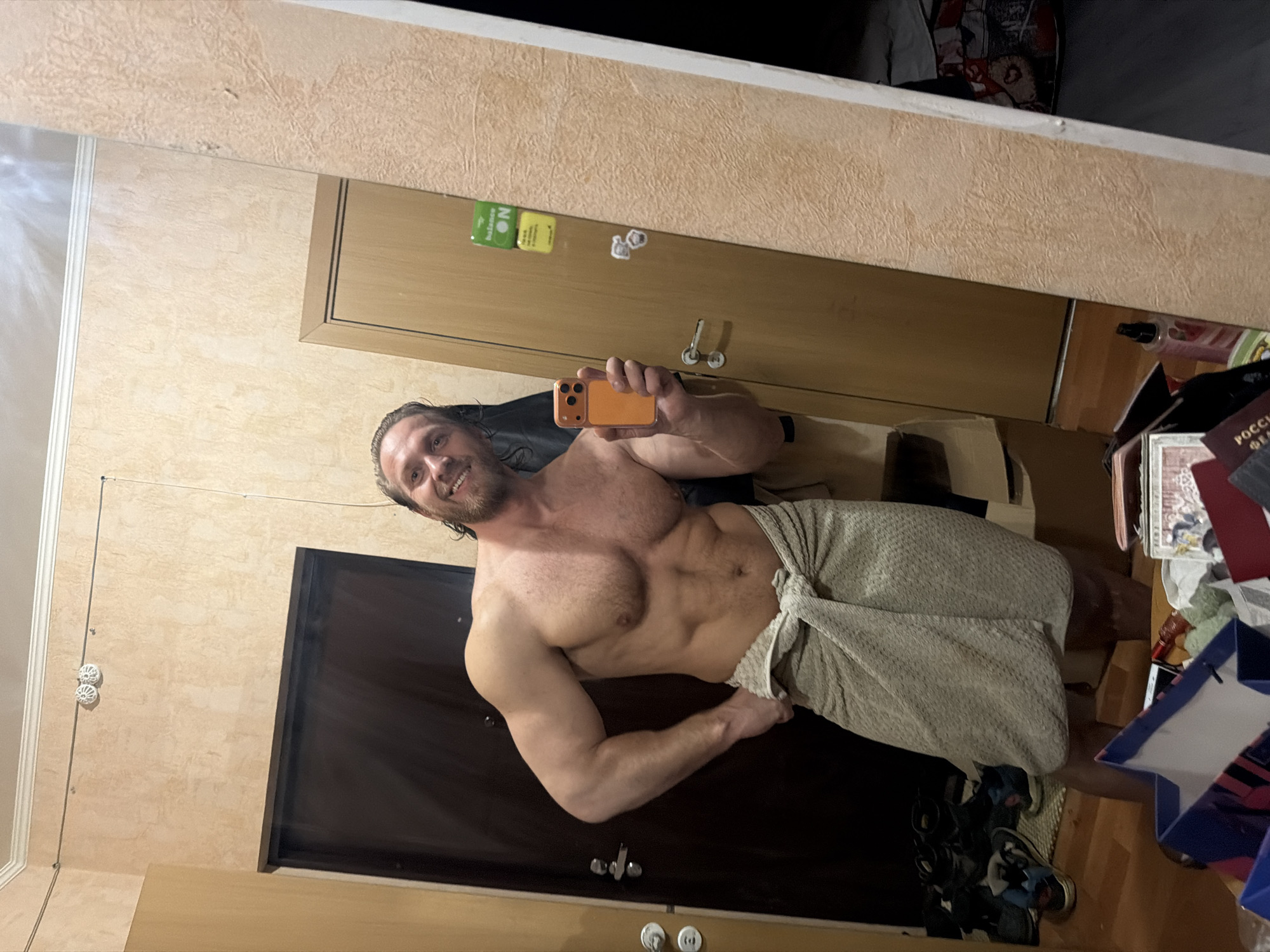 The image depicts a muscular adult male taking a mirror selfie in a residential interior, identified as Saint Petersburg, Russia. The man has wet, dark hair and a beard, is bare-chested, and wears a light-colored, textured towel wrapped around his waist. He smiles directly at the camera while holding an orange-cased smartphone in his right hand.
The environment features light beige, textured wallpaper and white crown molding along the ceiling, from which a thin dark cable with two white circular fixtures runs horizontally. In the reflection to the left of the man, a dark brown door with a silver handle is visible, with multiple pairs of shoes and a blue shopping bag on the wooden floor below. To the man's right, a light brown wooden door with a silver handle is visible, bearing a green "balance ON" sticker, a yellow sticker, and two small white decorative elements. On the floor in the bottom right, various items are scattered, including a bottle with pink liquid and a document bearing the text "РОССИЯ ФЕДЕРАЦИЯ."