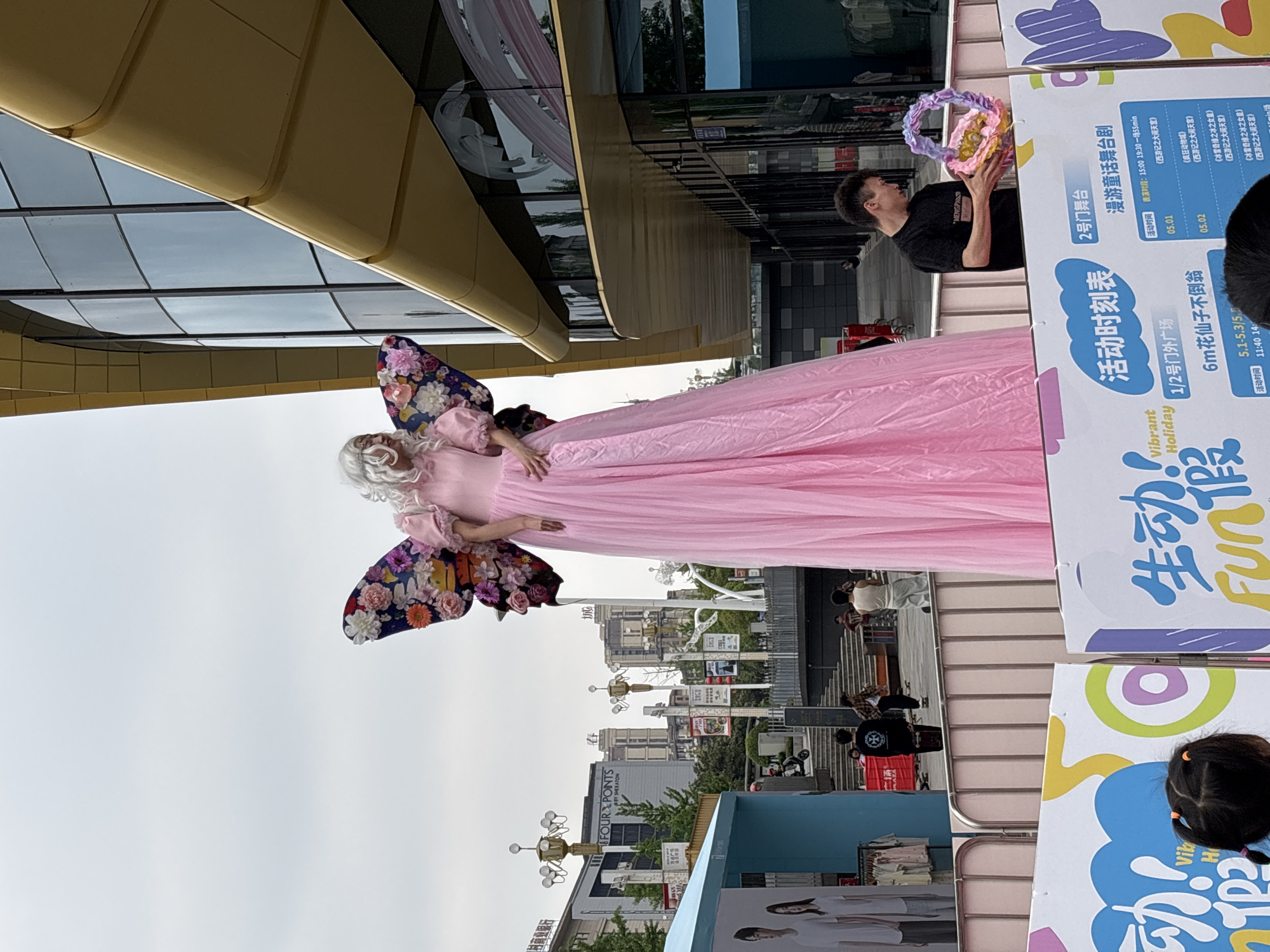 An outdoor scene in Jiangjin, China, captures a festive event in a commercial plaza under an overcast sky during daytime. The central subject is a performer costumed as a towering fairy or giant doll, adorned in a long, flowing pink dress, large butterfly wings decorated with vibrant floral patterns, and a white curly wig.
To the right, a man in a black t-shirt holds a colorful, circular, braided object, appearing to interact with or assist the performer. In the background, modern buildings with gold-toned panels and large glass windows rise, one clearly marked "FOUR POINTS BY SHERATON."
Prominent white and colorful signs with Chinese text advertise event details. They display "Vibrant Holiday" and "生动! Fun假" (Lively! Fun Holiday), along with an "活动时刻表" (Activity Schedule). Information includes a "2号门舞台" (Gate 2 Stage) hosting a "漫游童话舞台剧" (Wandering Fairy Tale Stage Play) from 15:00 to 19:30, featuring shows like *Zootopia*, *Journey to the West: Havoc in Heaven*, and *Frozen: Snow Queen* on specific dates (May 1st and 2nd). Another section highlights the "1/2号门外广场" (Gate 1/2 Outdoor Plaza) with a "6m花仙子不倒翁" (6m Fairy Lady Tumbler), referring to the large fairy performer, active from May 1st to 3rd, with a listed time around 11:40. A young child's head is partially visible at the bottom right, observing the scene. The overall impression is a lively public performance or promotional event at a shopping complex or hotel.