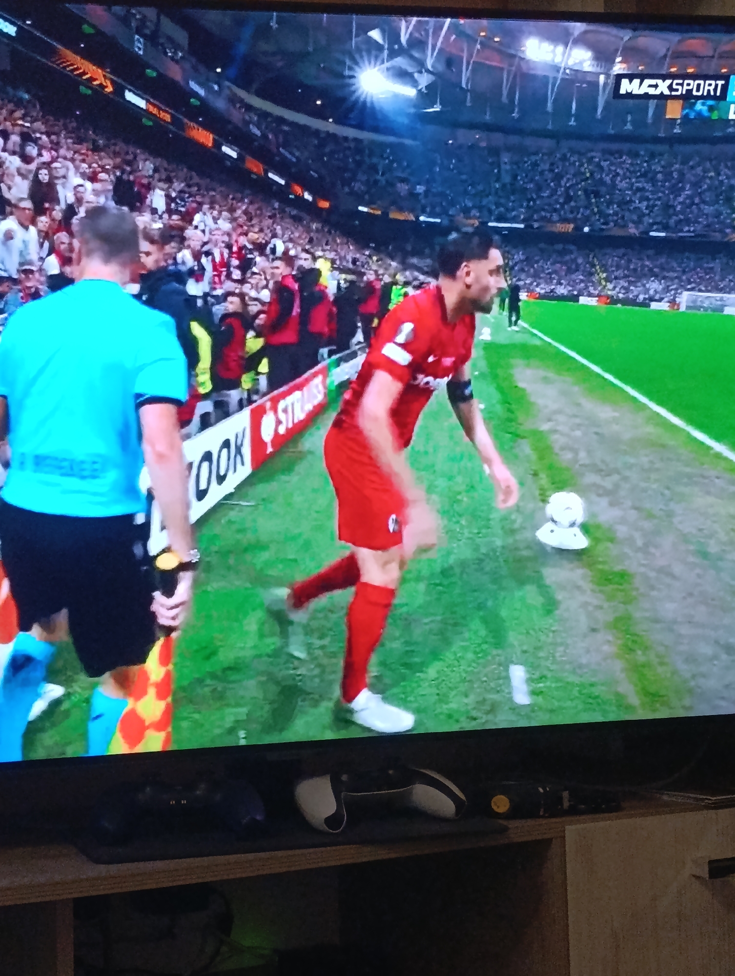 The image depicts a scene from a soccer match likely being viewed on a television. A soccer player in a red uniform stands on the sidelines of a green soccer field. A linesman is to his left. Beyond them, a stadium packed with spectators is visible. The television is on top of a wooden surface with gaming controllers. The match is being broadcasted on Max Sport. While the image is of a soccer match, there's no information to suggest it's taking place in Plovdiv, Bulgaria.