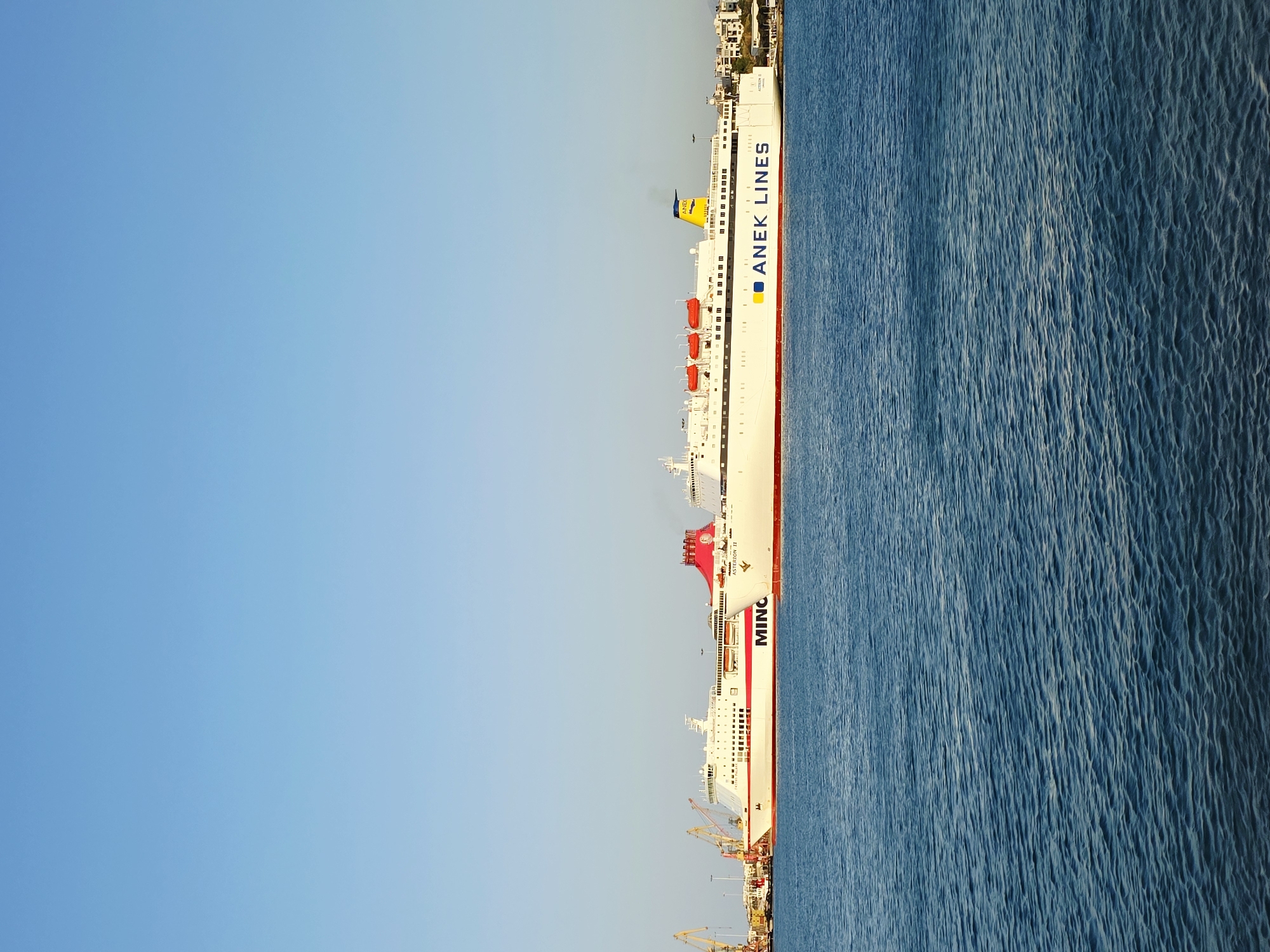 A large, white passenger ferry is docked in the harbor of Heraklion, Greece. The ferry, named "ASTERION II" and operated by "ANEK LINES," has a prominent red stripe along its hull and a yellow funnel with blue lettering. The ship is vast and spans a significant portion of the frame horizontally. In the background, on the left side of the image, there are several cranes and industrial structures, suggesting a port or shipyard. To the right of the ferry, a shoreline with buildings and some vegetation is visible. The sea in the foreground is calm and a deep blue, with gentle ripples on its surface. The sky above is clear and a light blue, indicating a sunny day. There are no people clearly visible on the ferry or in the surrounding area. The overall scene depicts a typical port environment with a prominent ferry.