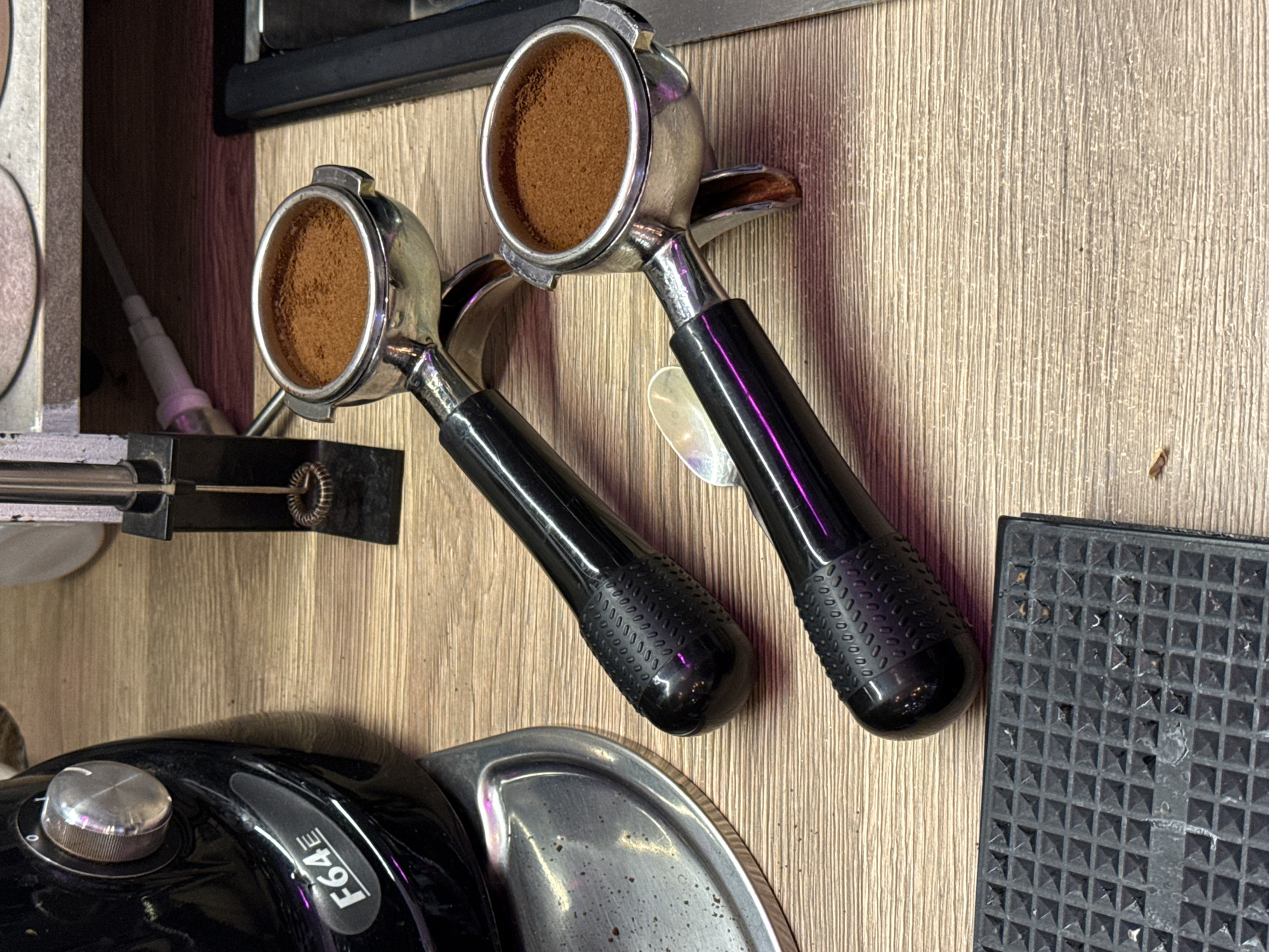 An overhead close-up shot captures two portafilters filled with ground coffee, resting on a light-colored wooden countertop. The portafilters have black handles with textured grips. To the left, a portion of a black coffee grinder is visible, with a silver dial and a label reading "F64E". In the background, a metal frothing wand stands upright, attached to a black base. To the far right, a metallic surface suggests a sink or a part of the coffee machine. The lighting in the scene is slightly dim, with purple reflections visible on the portafilter handles. The image does not provide any information related to Moscow, Russia.