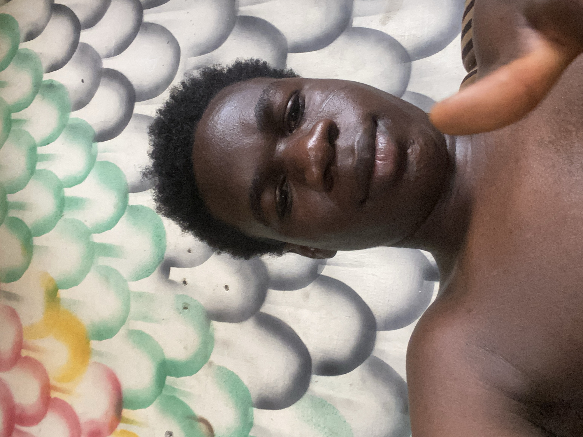 A close-up, rotated image features a dark-skinned person with short, dark hair. The individual's face is centered in the frame. A portion of their bare shoulder and upper chest is also visible. A hand is partially visible, blurry, and out of focus on the right side of the image. The background consists of a wall with a painted design resembling scales in shades of white, grey, green, yellow, and pink. The location is Mararaba, Nigeria.