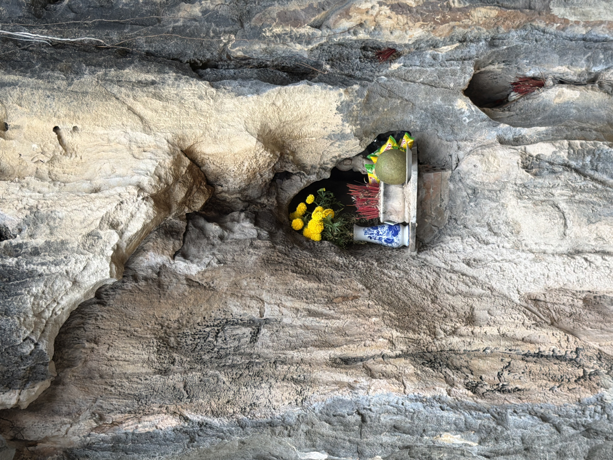 The image shows a small shrine carved into a rock face at Marble Mountains in Da Nang, Vietnam. The shrine is located in a natural crevice in the textured, grey and tan rock. Inside the crevice, a white ceramic vase with blue floral patterns holds a bouquet of bright yellow flowers. To the right of the vase, a collection of red incense sticks are bundled together in a pot. In front of the incense, a round, green fruit rests on a small plate, and two yellow and green wrappers are visible behind it. A small, light-colored statue, possibly of a deity, is partially visible behind the incense and fruit. Further down and to the right, another small niche in the rock contains a bundle of red incense sticks. The rock face displays natural erosion and varying shades of grey, brown, and tan.