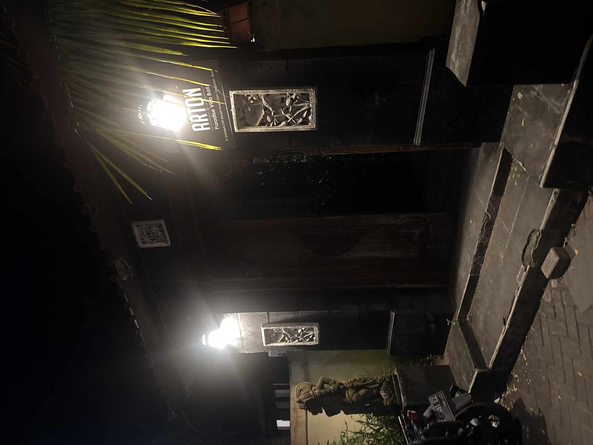 Here is a description of the image:
The image captures a nighttime view of the entrance to "Arton Paradise Villa Bali," located in Ungasan, Bali, Indonesia. A sign illuminated by a bright light displays the establishment's name and address: "Jl. BKR Bali Cliff, Banjar Anges Sari, Ungasan-Bali." The entrance features decorative elements such as carved stone panels and a statue. Part of a motorcycle is visible at the bottom of the frame, parked in front of the entrance. The scene is dimly lit, with artificial light sources illuminating key features of the building and sign.