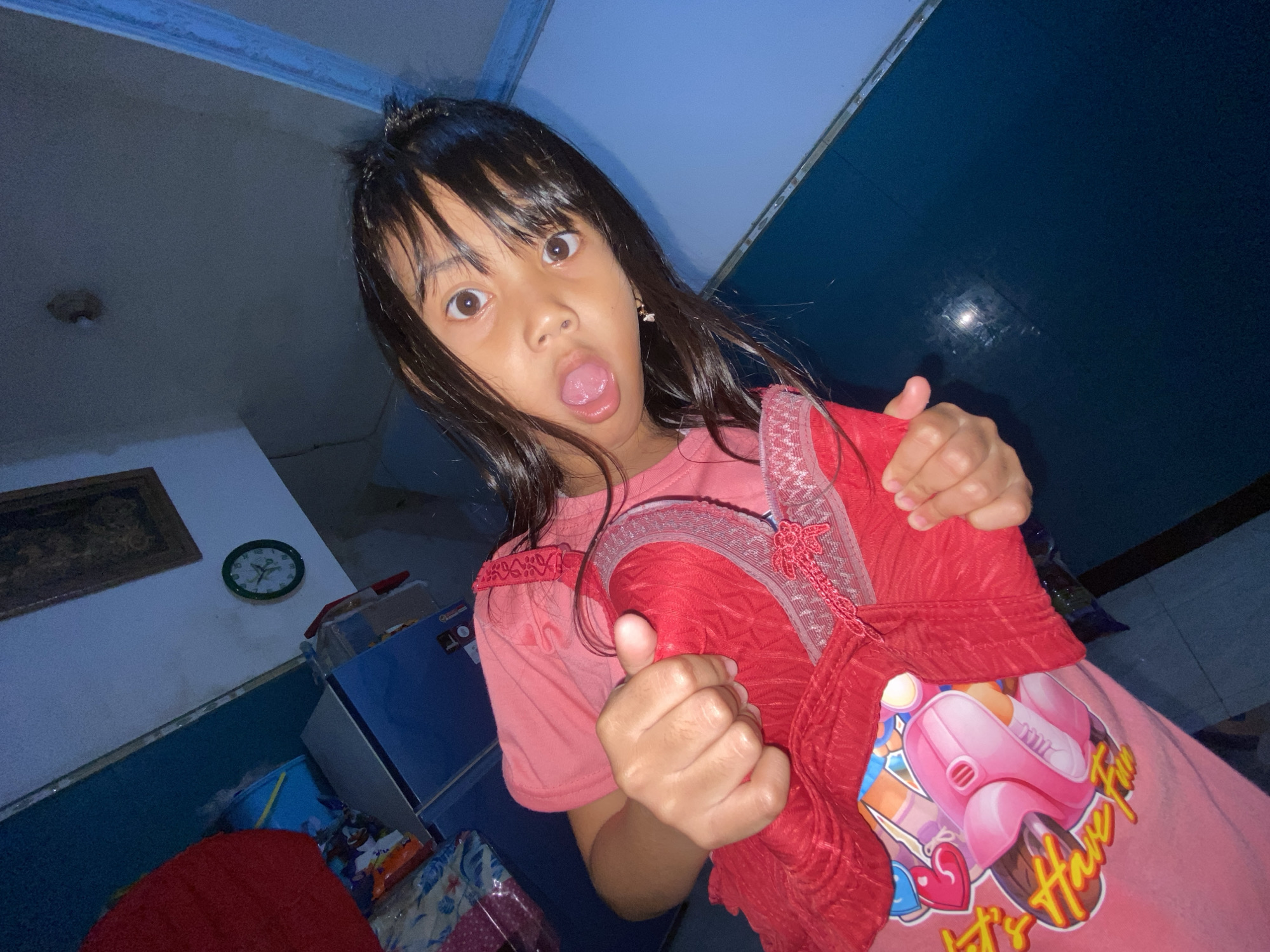 A young girl with black hair is indoors with her tongue sticking out, holding a red garment. She is wearing a pink t-shirt with a graphic design on it. The background includes a clock, a framed picture, and a blue refrigerator. The location is possibly Gelam, Indonesia.