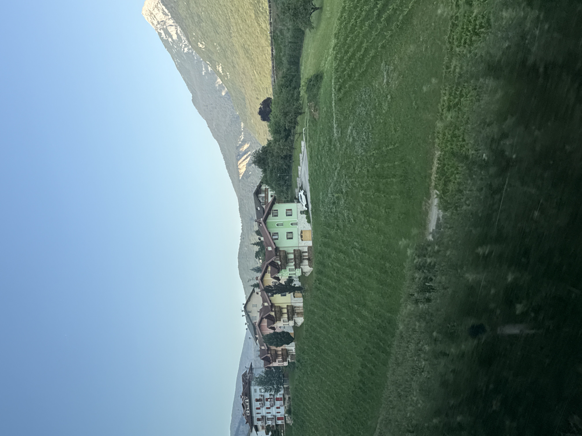 A serene panorama captures the charming village of Spormaggiore, Italy, nestled against the backdrop of majestic mountains. The image showcases a cluster of colorful houses, their facades painted in vibrant hues of green, yellow, and pink, adorned with balconies and intricate architectural details. These residences are strategically positioned on a gentle slope, overlooking a verdant expanse of terraced vineyards that cascade down the hillside. A pristine white car is parked on the paved area adjacent to one of the houses, hinting at a tranquil, leisurely lifestyle. The foreground is dominated by the lush greenery of the vineyards, their rows clearly defined, adding a touch of natural beauty to the scene. The sky above is a clear, unblemished blue, transitioning into a soft gradient towards the horizon. The mountains, rugged and imposing, stand tall in the distance, their peaks kissed by the warm sunlight, creating a striking contrast with the serene village below. The overall impression is one of peace and tranquility, a harmonious blend of human settlement and natural splendor.