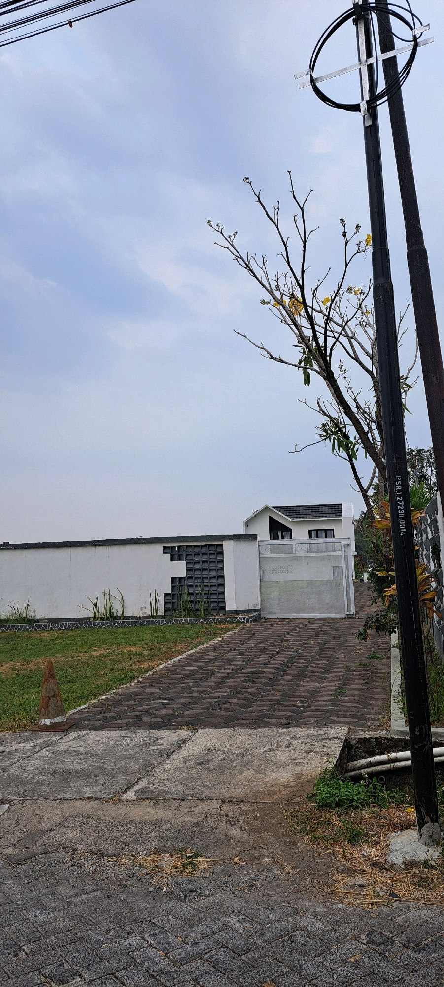 The image depicts a view of a modern house with a white facade, a gated driveway, and a surrounding yard. The scene is set in Ketanireng, Indonesia.
In the foreground, a paved driveway made of dark interlocking bricks leads towards a silver-colored gate. The driveway is partially cracked and worn in places, with some debris and dry grass visible. A concrete path runs alongside the driveway. To the left of the driveway, a patch of green grass is visible, marked by a conical traffic marker.
A long, white wall with a decorative pattern of dark glass blocks extends along the left side of the property. Some slender green plants grow along the base of this wall.
The house itself is visible in the mid-ground. It features a white exterior, a dark-tiled roof, and prominent angled architectural elements. A white gate separates the driveway from the house.
To the right of the scene, a tall, dark utility pole with visible wires and a metallic crossbar stands prominently. A tree with bare branches and a few yellow flowers is situated between the utility pole and the house. The sky is overcast with a pale blue hue. The overall environment suggests a residential area with a focus on modern architecture.