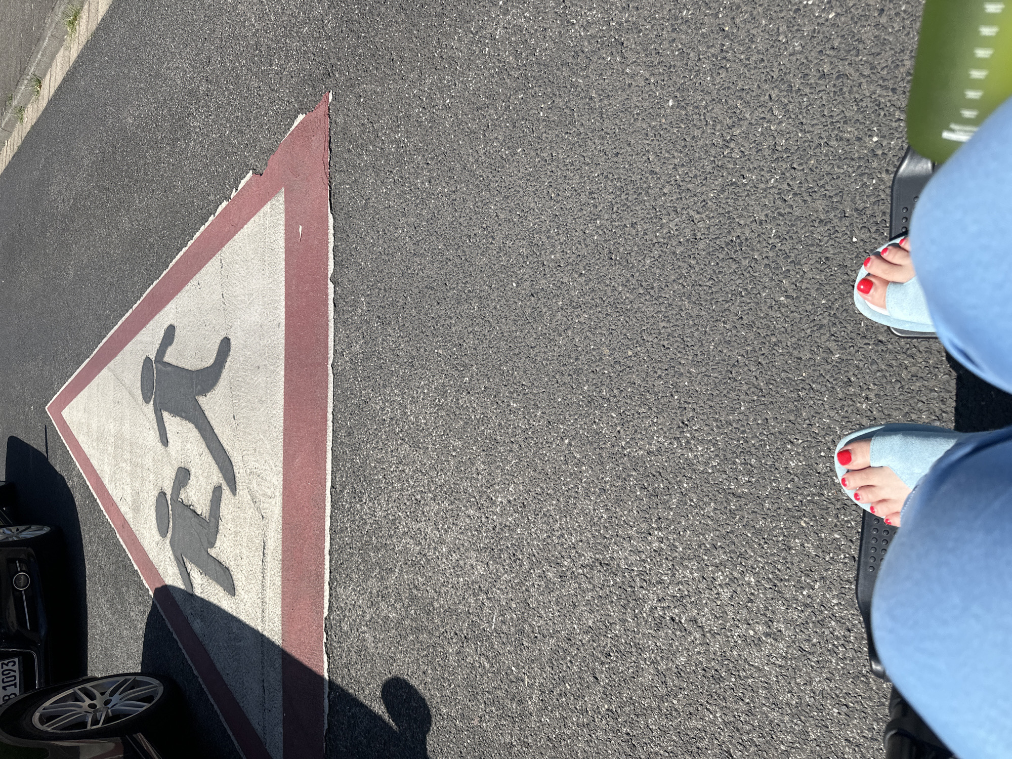 This is a low-angle, eye-level shot of asphalt road with a caution sign painted on it. The sign is a white triangle with a red border and depicts two dark grey human figures, one behind the other, in a running stance. The sign is faded in places, especially the white inner triangle. To the left of the sign, the front wheel and part of the body of a dark colored car is visible. The car has a license plate that reads "B 1093". In the foreground, the legs and feet of a person wearing light blue pants and light blue open-toed shoes are visible. The toes of the person are painted with red nail polish. The person is sitting in a wheelchair. The scene is lit by bright sunlight, casting shadows. The location is Krefeld, Germany.