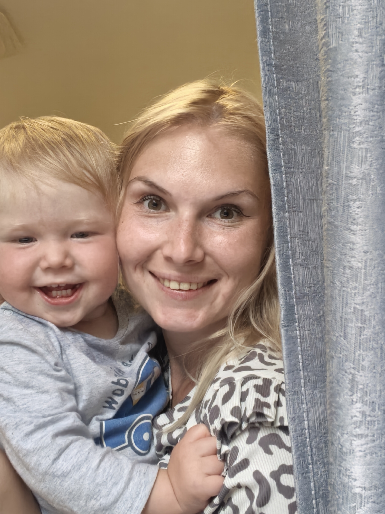 A smiling blonde woman holds a laughing blonde toddler. The woman is on the right, and the toddler is on the left. The woman is wearing a gray and white leopard-print top. The toddler is wearing a gray long-sleeved shirt. Part of a blue curtain is visible on the right side of the image. The background is a blurred yellow wall. The image appears to have been taken indoors.