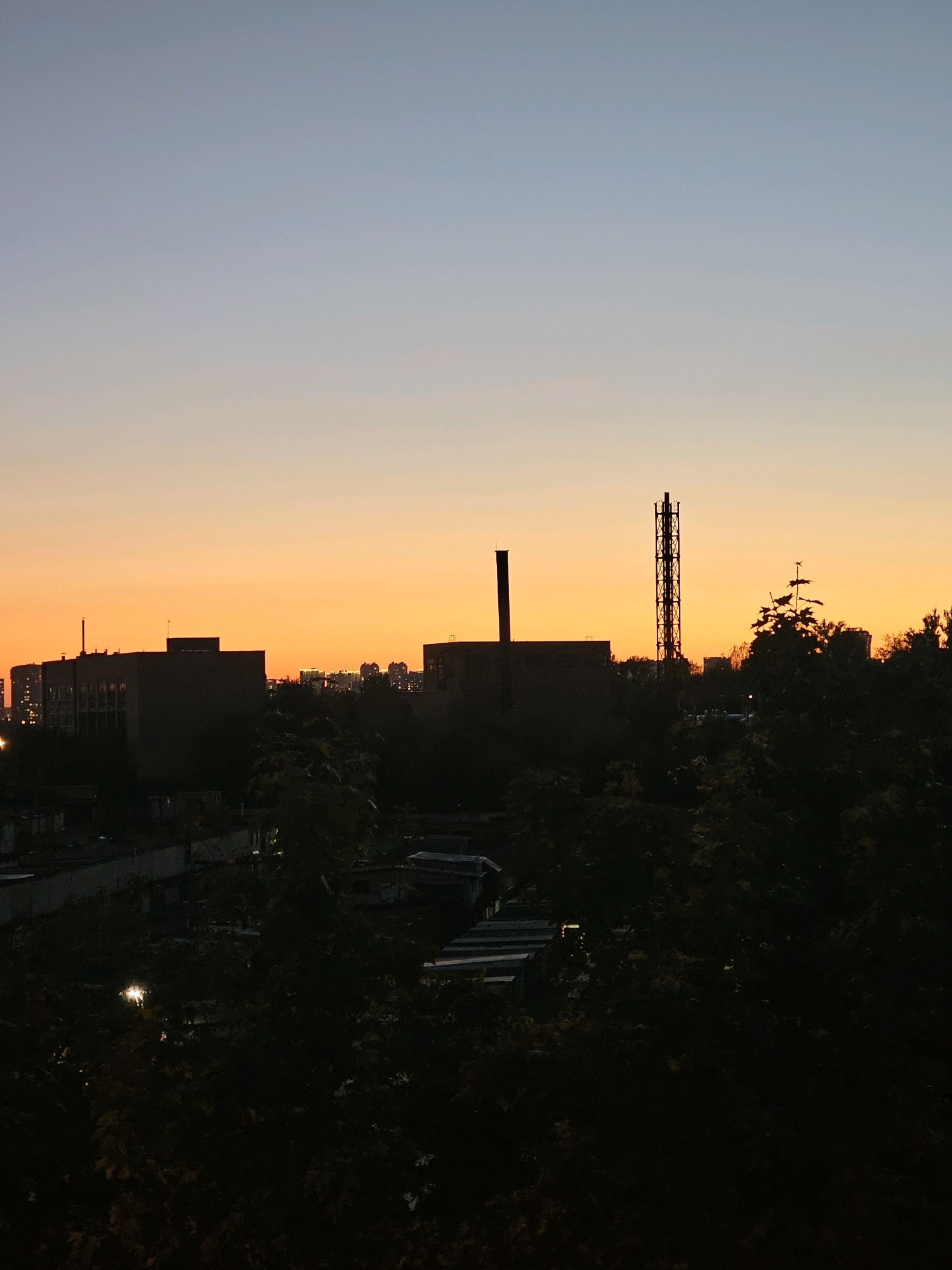 This is a photograph of a silhouetted cityscape at dusk, taken from a high vantage point. The sky transitions from a pale blue at the top to a vibrant orange and yellow gradient near the horizon. In the foreground, dark foliage obscures much of the lower portion of the image, with some glimpses of low-rise buildings and structures.
Prominently featured against the sky are several tall, dark silhouettes of industrial or urban structures. To the left, a rectangular building with several visible windows is silhouetted. To the right of this, a tall, lattice-like industrial tower stands out. Between these structures, more dark trees and some less defined buildings are visible. In the distance, a faint outline of more city buildings can be seen.
The scene appears to be an urban environment, possibly an industrial or residential area on the outskirts of a city. Based on the provided context, the location is identified as the city of Khimki, Russia. There are no people visible in the frame, and the focus is on the architecture and the atmospheric conditions of the sunset. The lighting is low, with the setting sun casting a warm glow on the horizon and creating strong silhouettes of the objects in the foreground and midground.